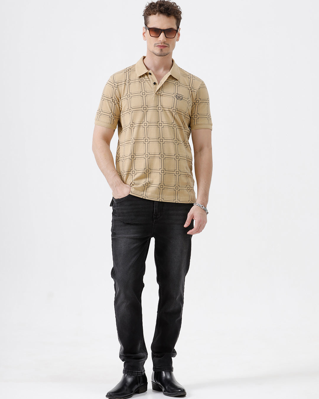 A man wearing a beige short-sleeve polo shirt with a brown grid pattern, paired with black jeans, standing against a plain white studio background.