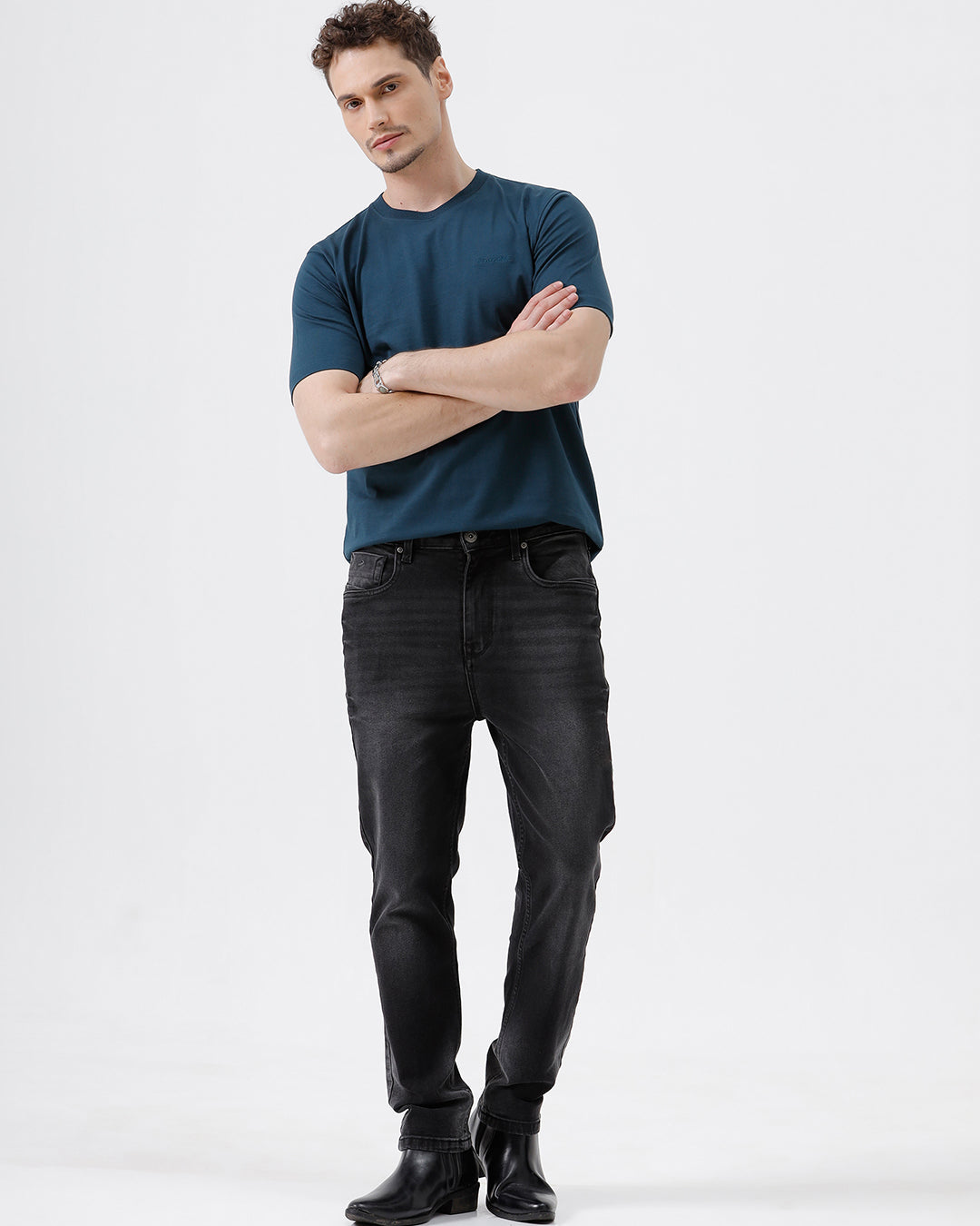 A person wearing slim-fit black denim jeans with a faded wash, paired with a dark blue t-shirt and black leather boots, shown from the waist down on a light gray studio background.