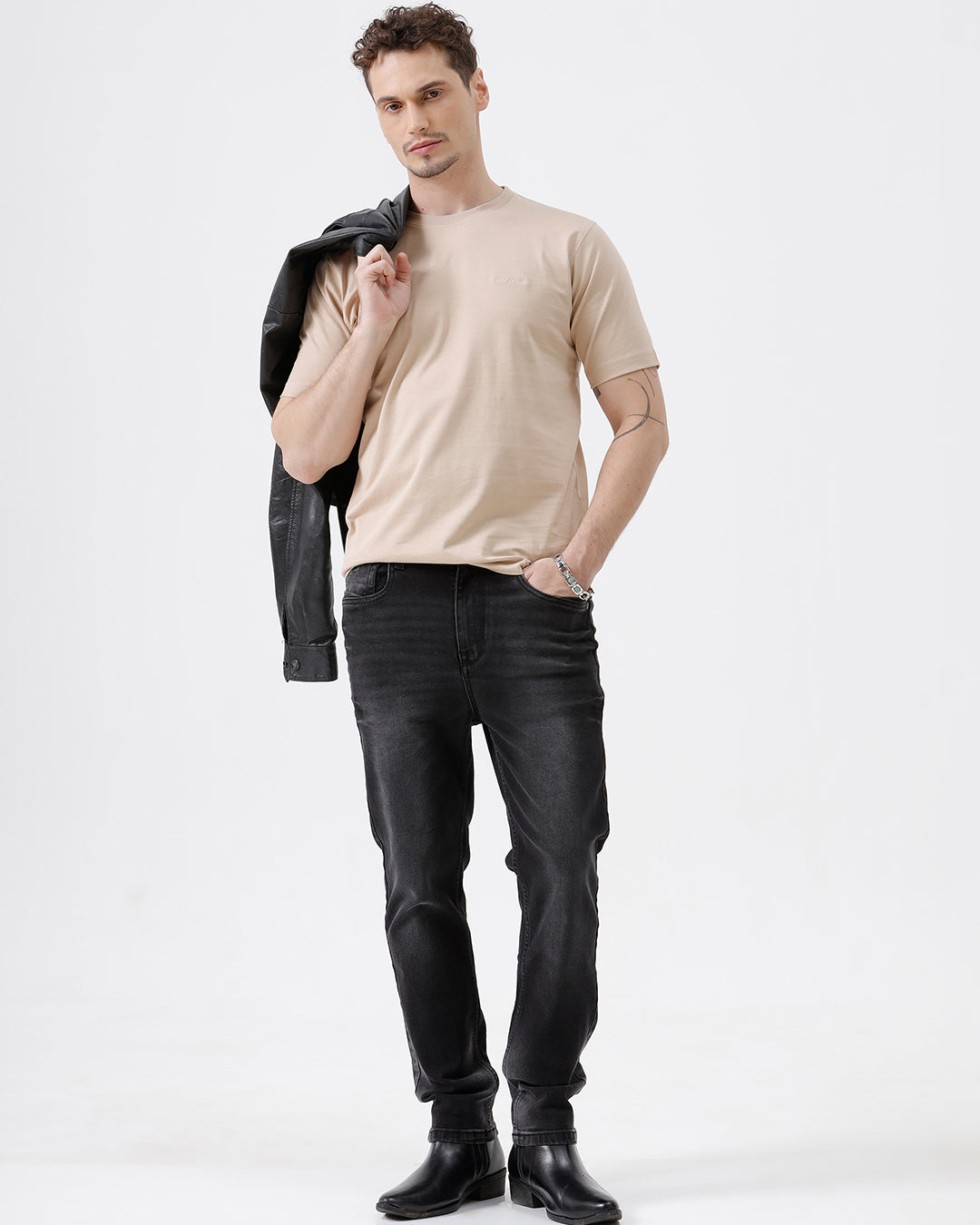 A man wearing a beige short-sleeve crewneck t-shirt with a subtle tonal logo on the left chest, paired with black jeans, standing against a light gray studio background.