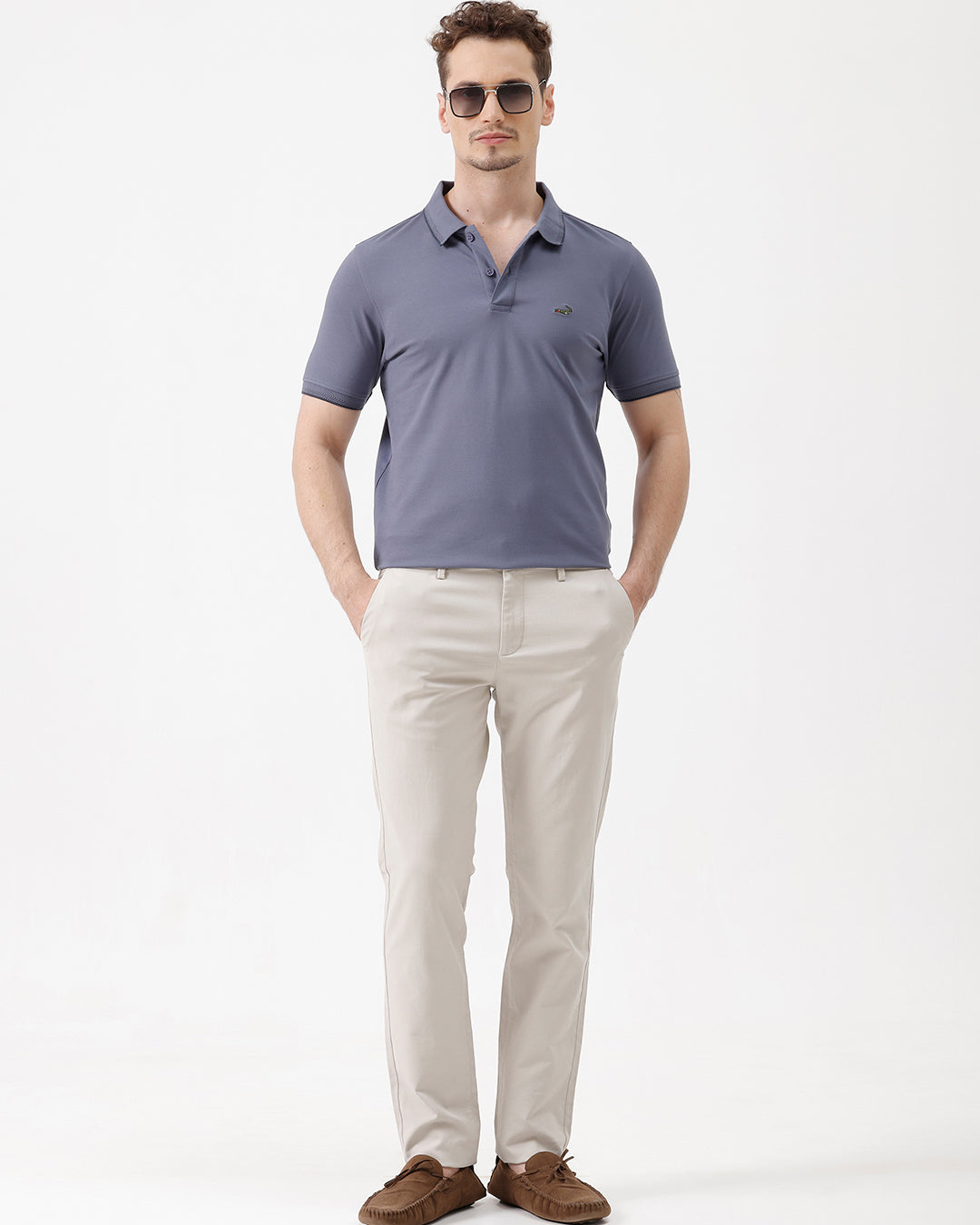 A man wearing a slate blue polo shirt and beige trousers, standing against a plain white background.