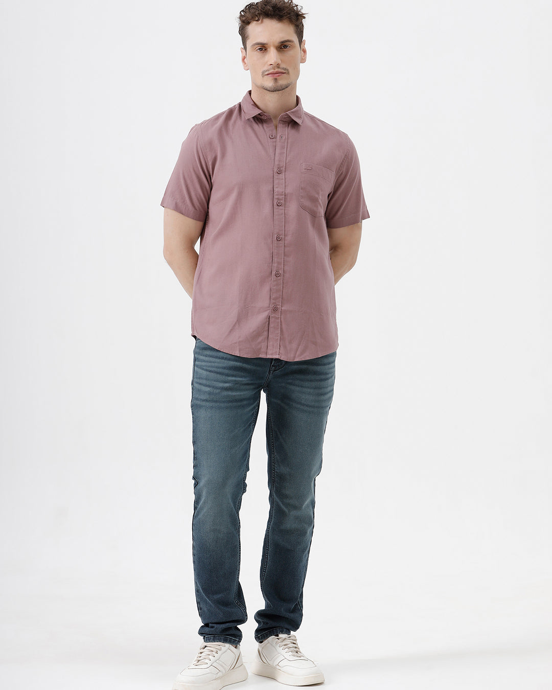 A man wearing a short-sleeve button-up shirt in dusty rose pink with dark blue jeans, standing against a plain white studio background.