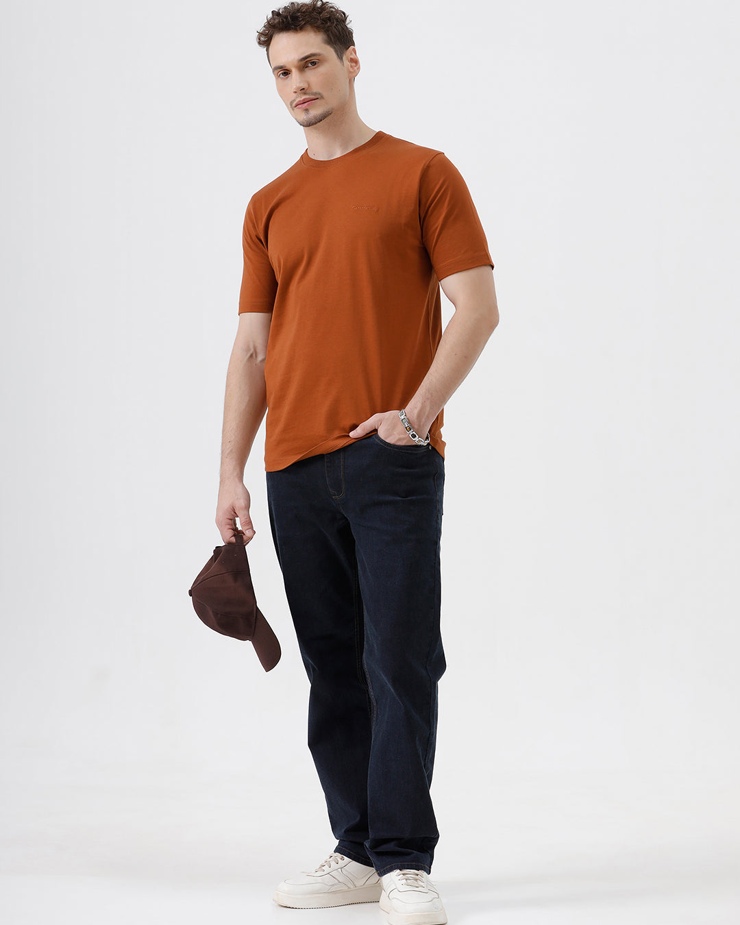 A man wearing a rust-orange crew-neck t-shirt and dark blue jeans, standing against a light gray studio background.