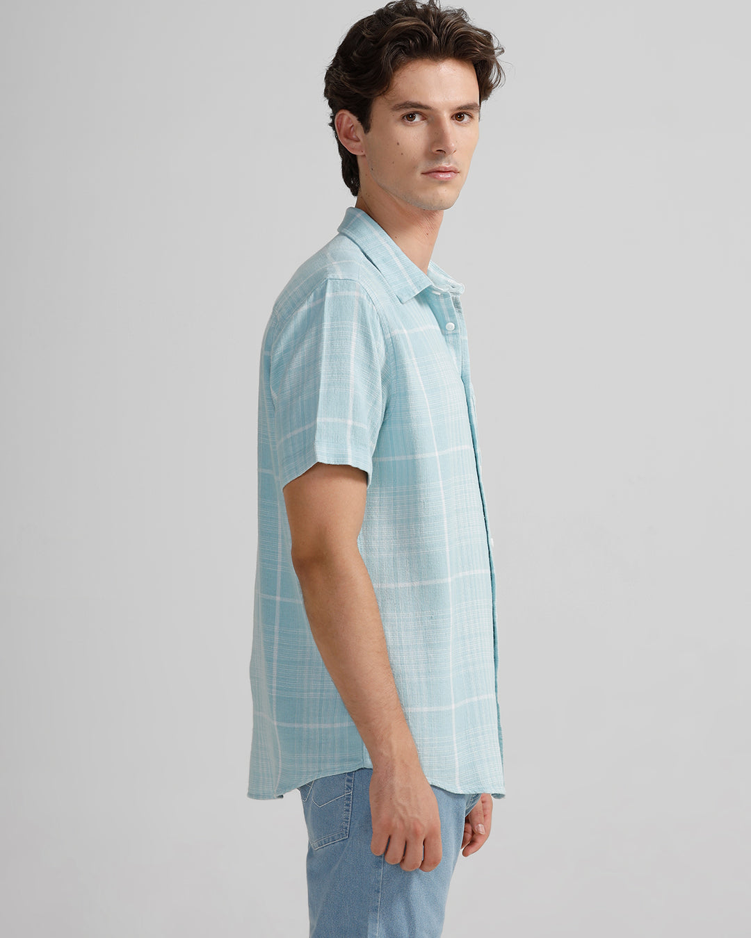 Man wearing a light blue checkered shirt and jeans against a plain background.