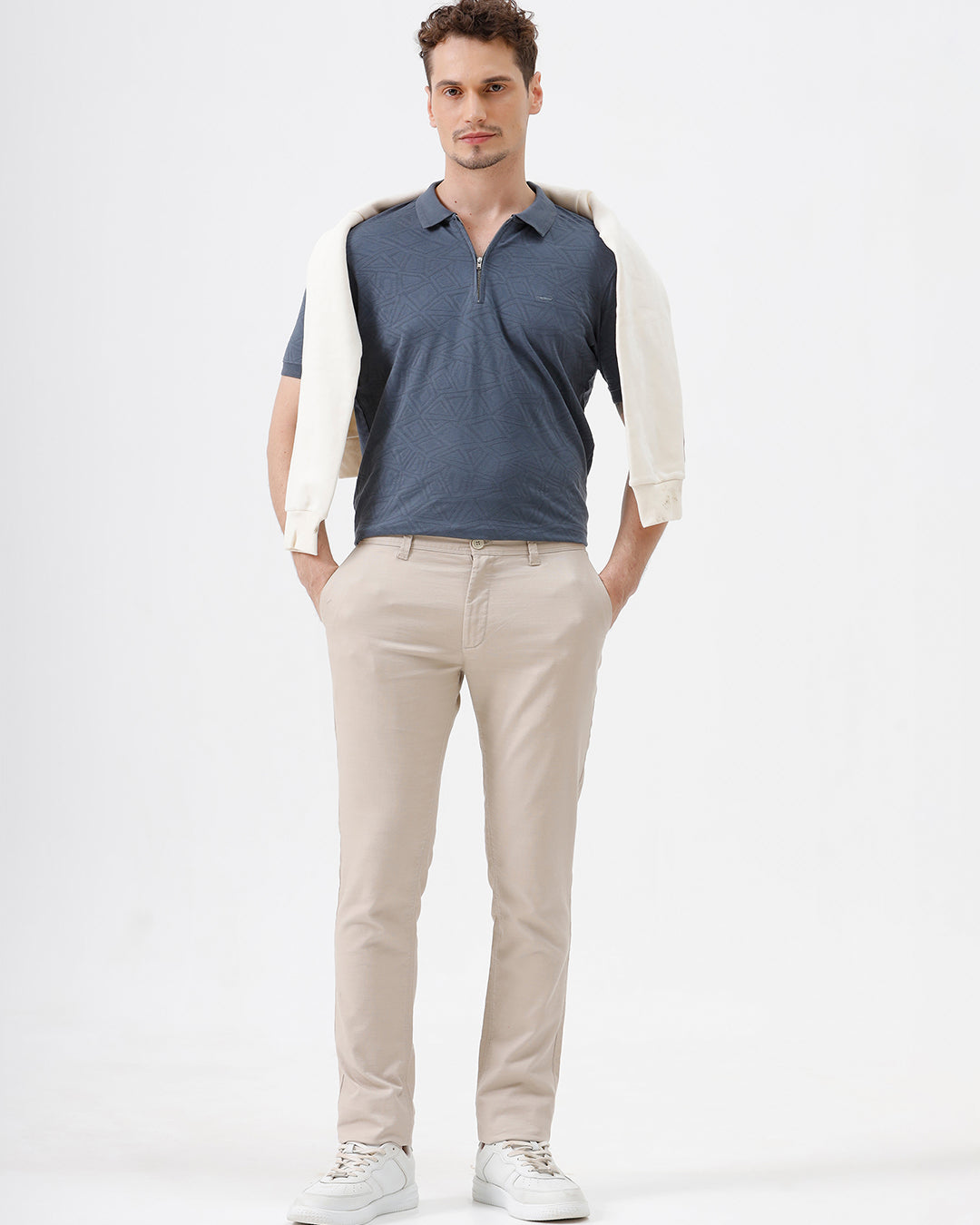 A person wearing slim-fit beige chinos with a blue patterned t-shirt and white sneakers, standing against a white studio background.