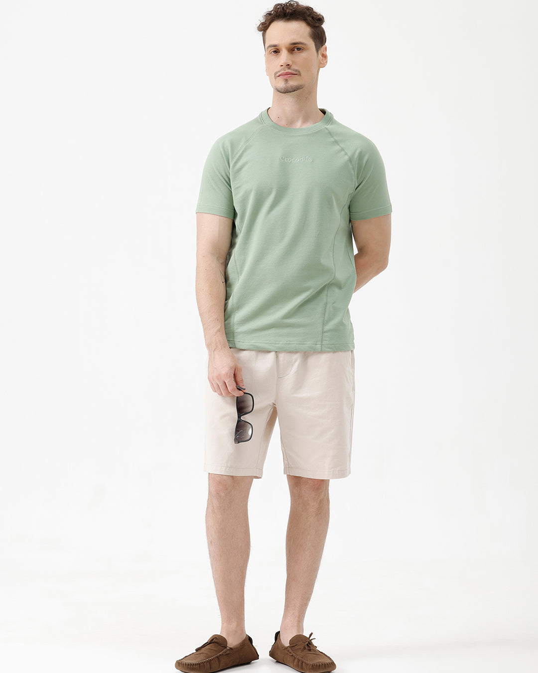 A man wearing a light sage green athletic t-shirt with a small tonal logo on the chest, paired with beige pants, standing against a plain white studio background.