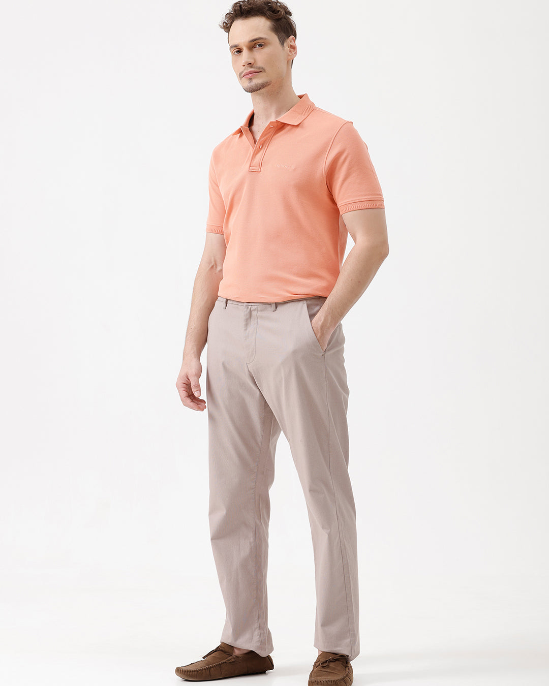 A man wearing a peach-colored short-sleeve polo shirt and beige trousers, standing against a plain white studio background.