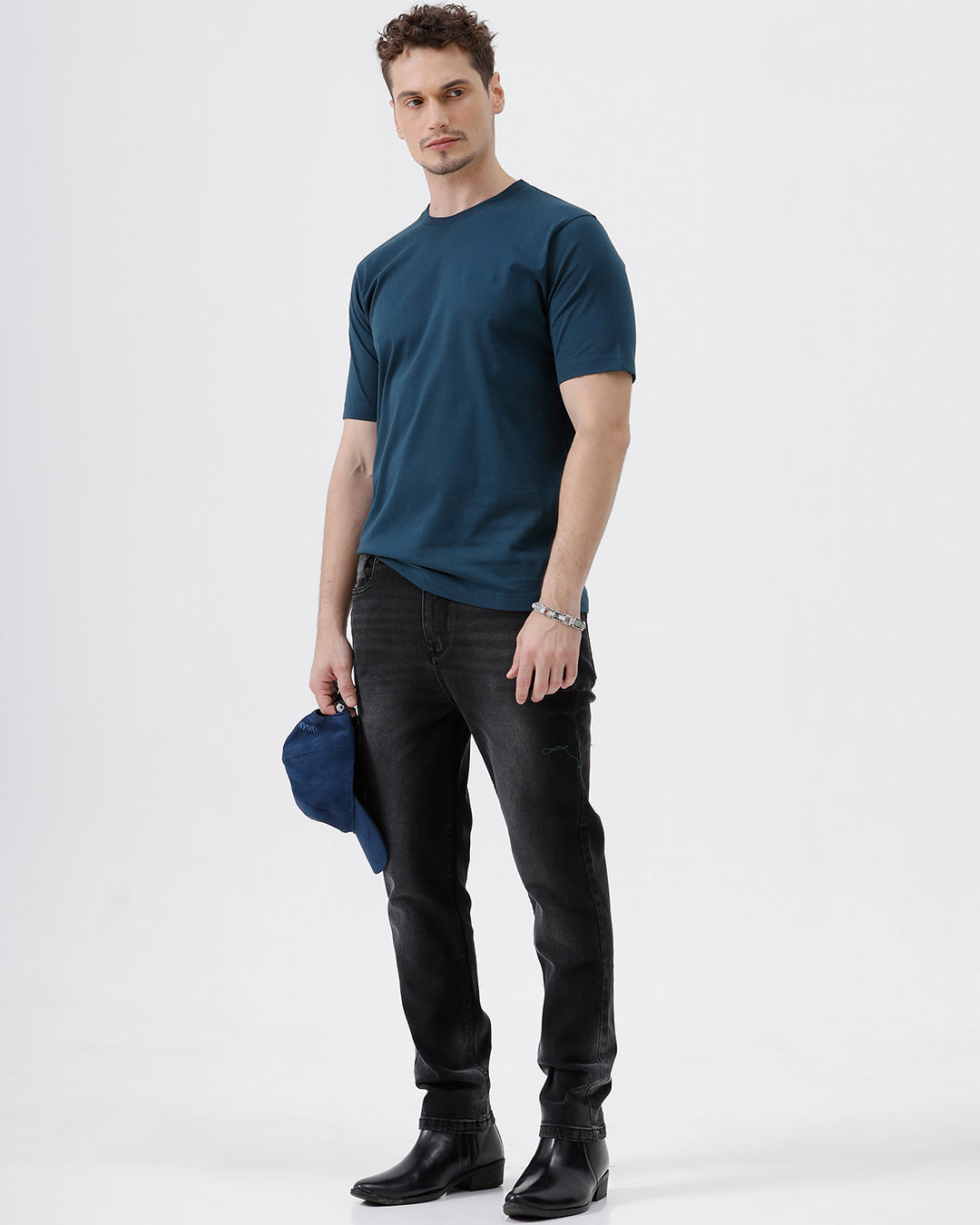 A man wearing a dark navy blue crew-neck t-shirt and black jeans, standing against a light gray studio background.