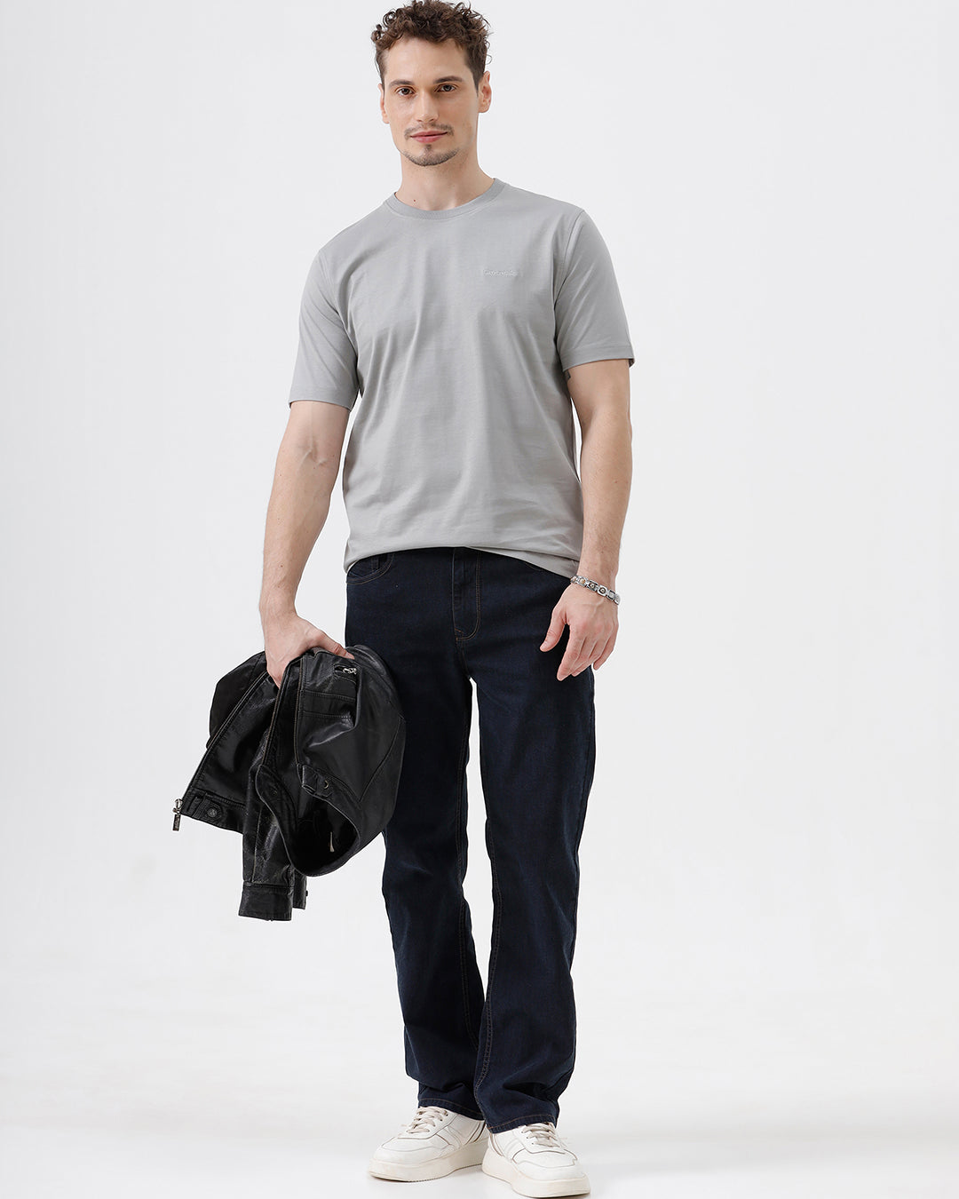 A man wearing a light gray short-sleeve crewneck t-shirt with a subtle tonal chest logo, paired with dark jeans, standing against a light gray studio background.