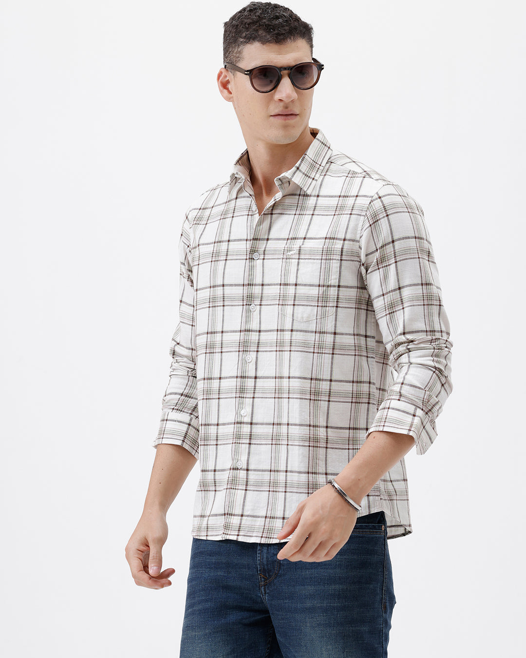 A man wearing a long-sleeve plaid button-up shirt in beige and brown with dark blue jeans, standing against a plain white studio background.