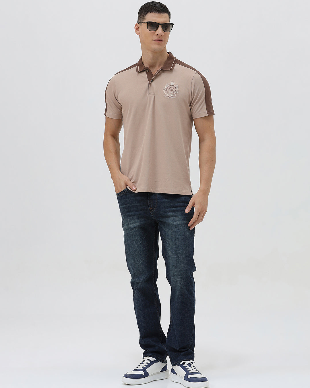 A man modeling a short-sleeve polo shirt in beige with brown shoulder panels and collar trim, paired with dark blue jeans, against a light gray studio background.