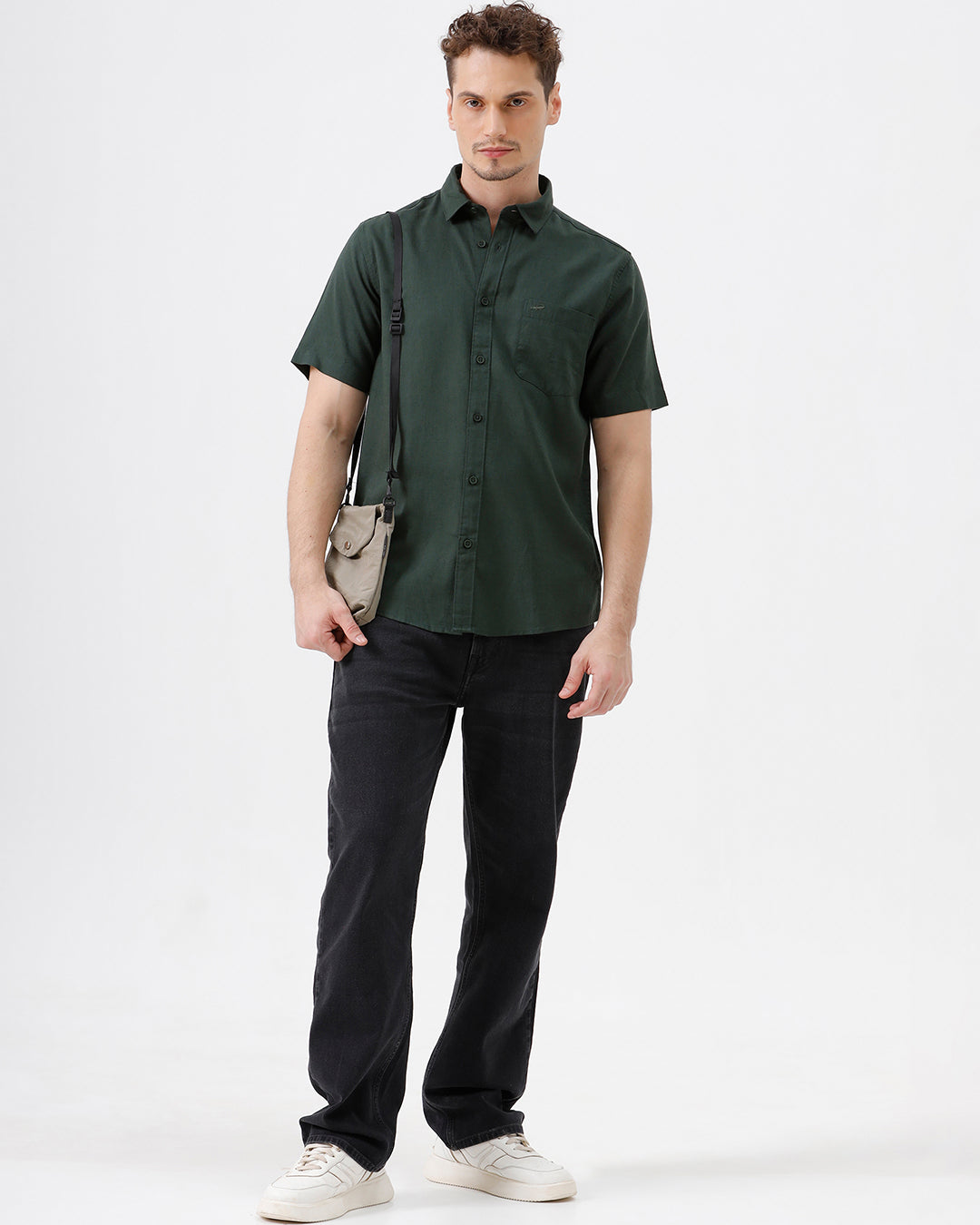 A man wearing a dark forest green short-sleeve button-up shirt with a chest pocket, paired with black jeans, standing against a plain white studio background.