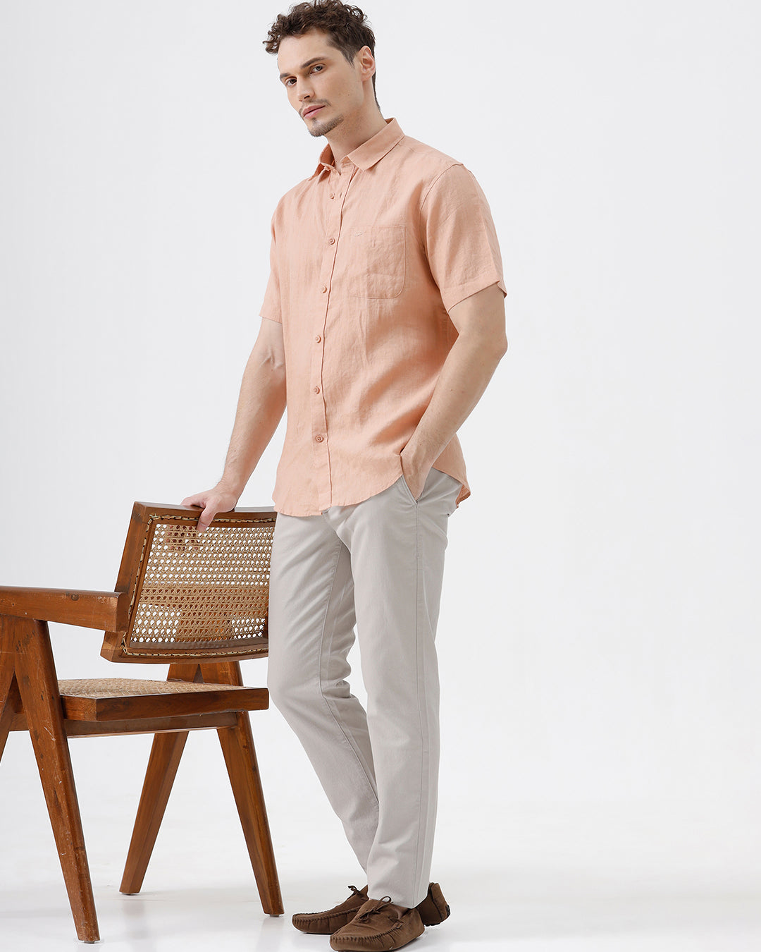 A man wearing a short-sleeve linen button-up shirt in a light peach-pink color, paired with light gray trousers, standing against a plain light gray background.