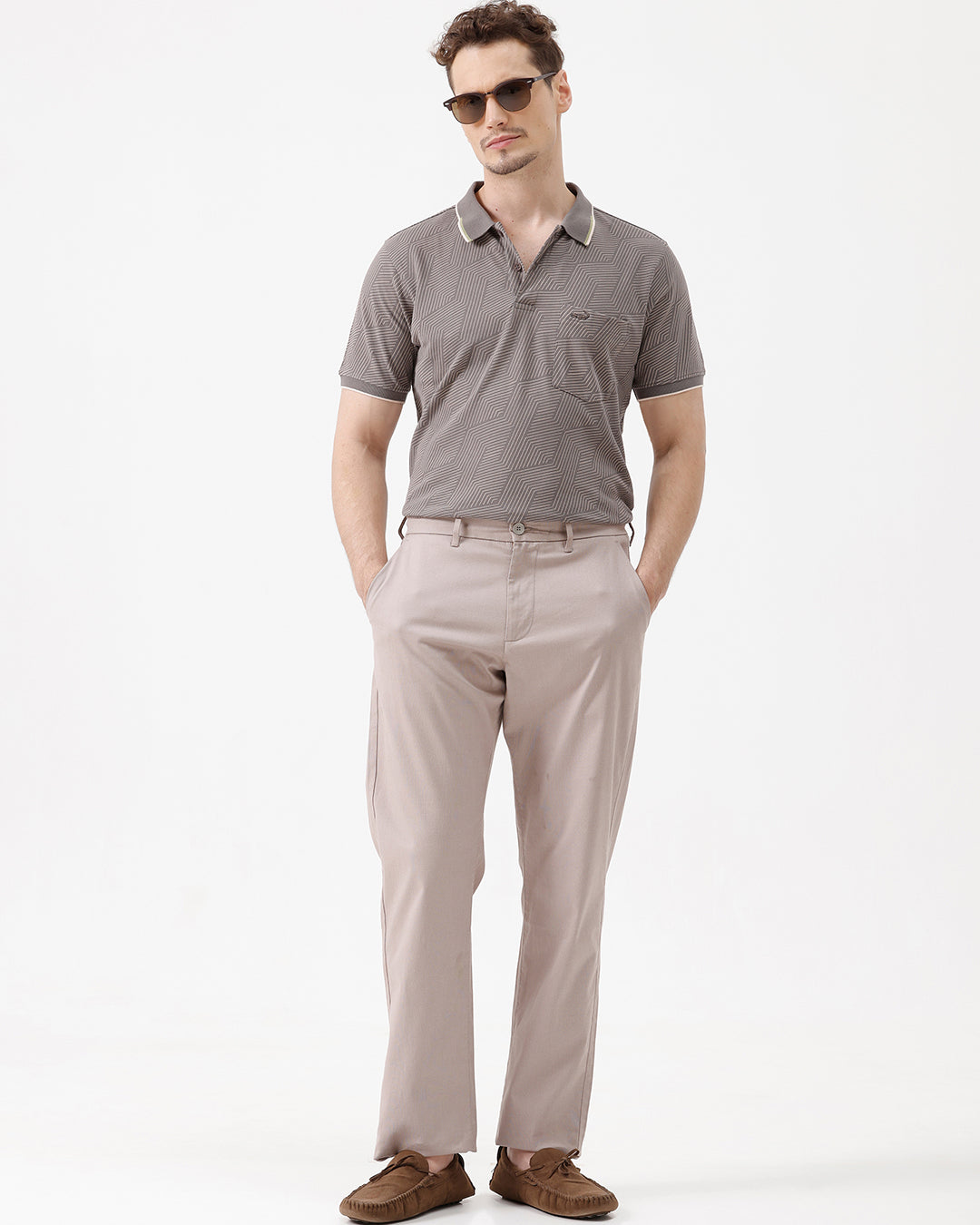 A man wearing a short-sleeve polo shirt with a gray and white geometric pattern, paired with beige trousers, standing against a plain white studio background.