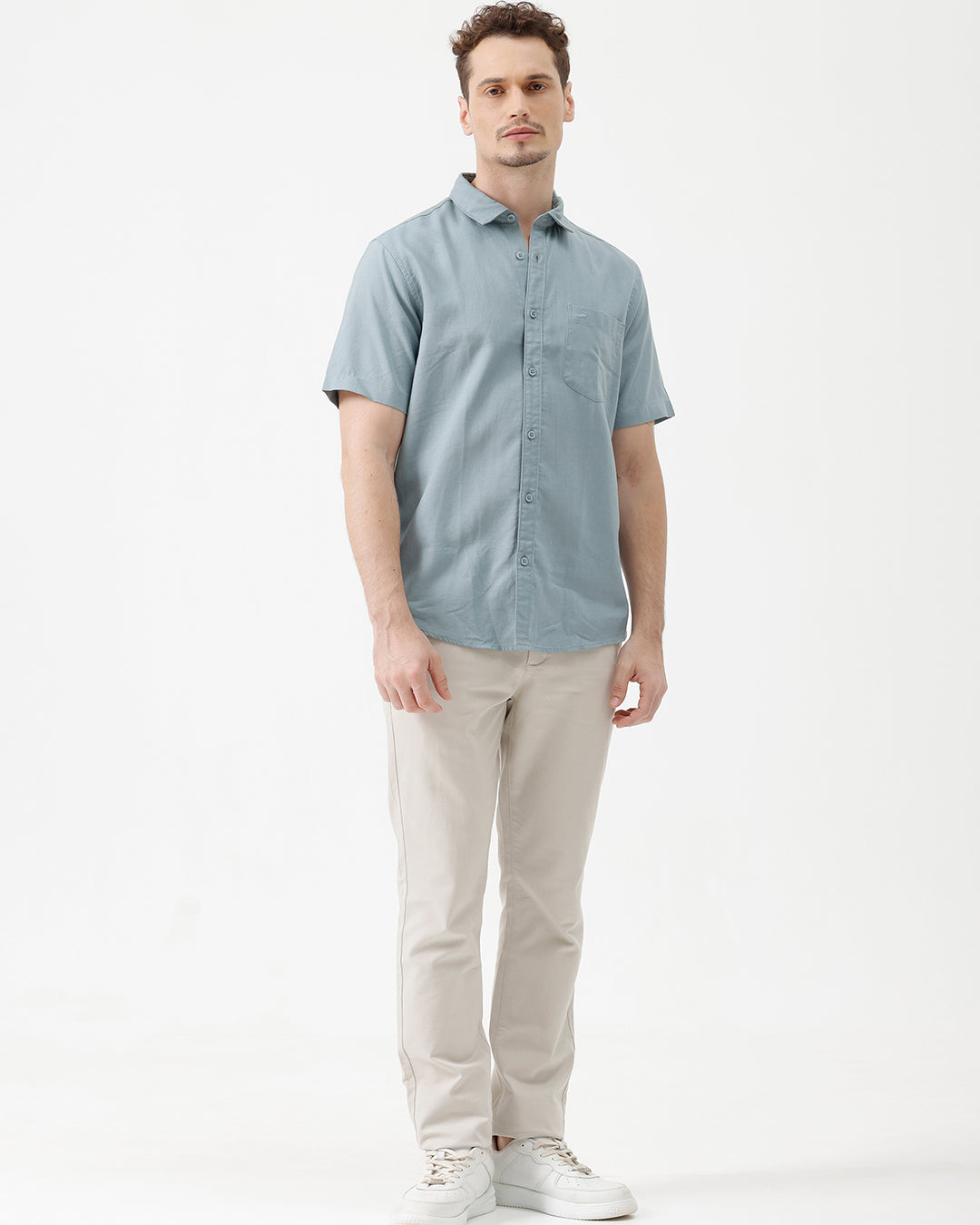 A man wearing a light blue short-sleeve button-up shirt and beige trousers, standing against a plain white studio background.