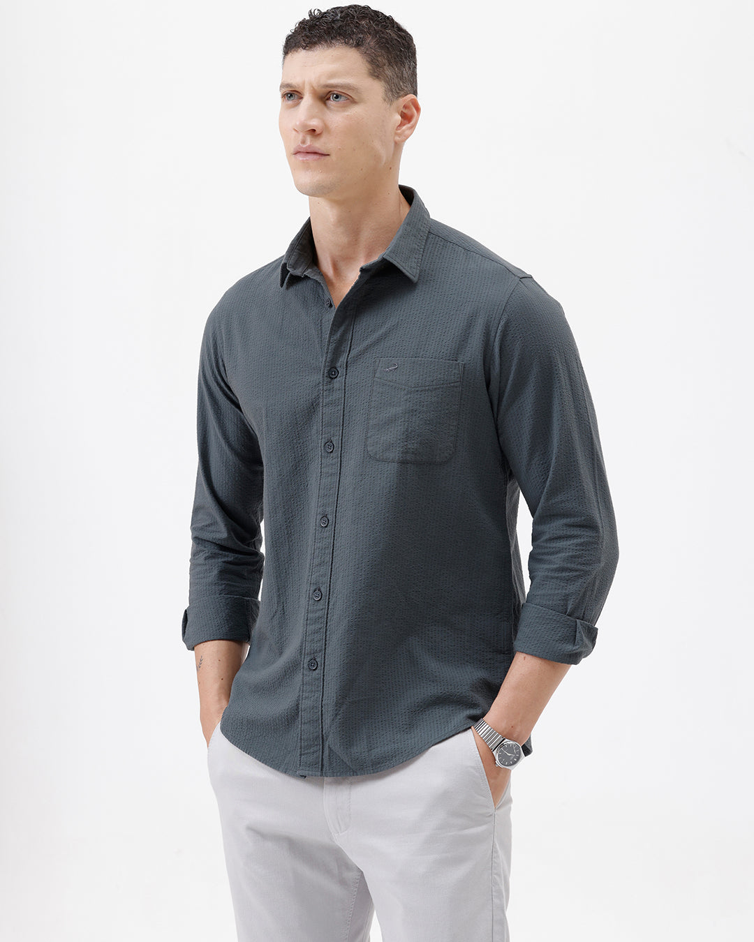 A man wearing a dark charcoal gray long-sleeve button-down shirt with a chest pocket, paired with light gray trousers, standing against a plain white studio background.