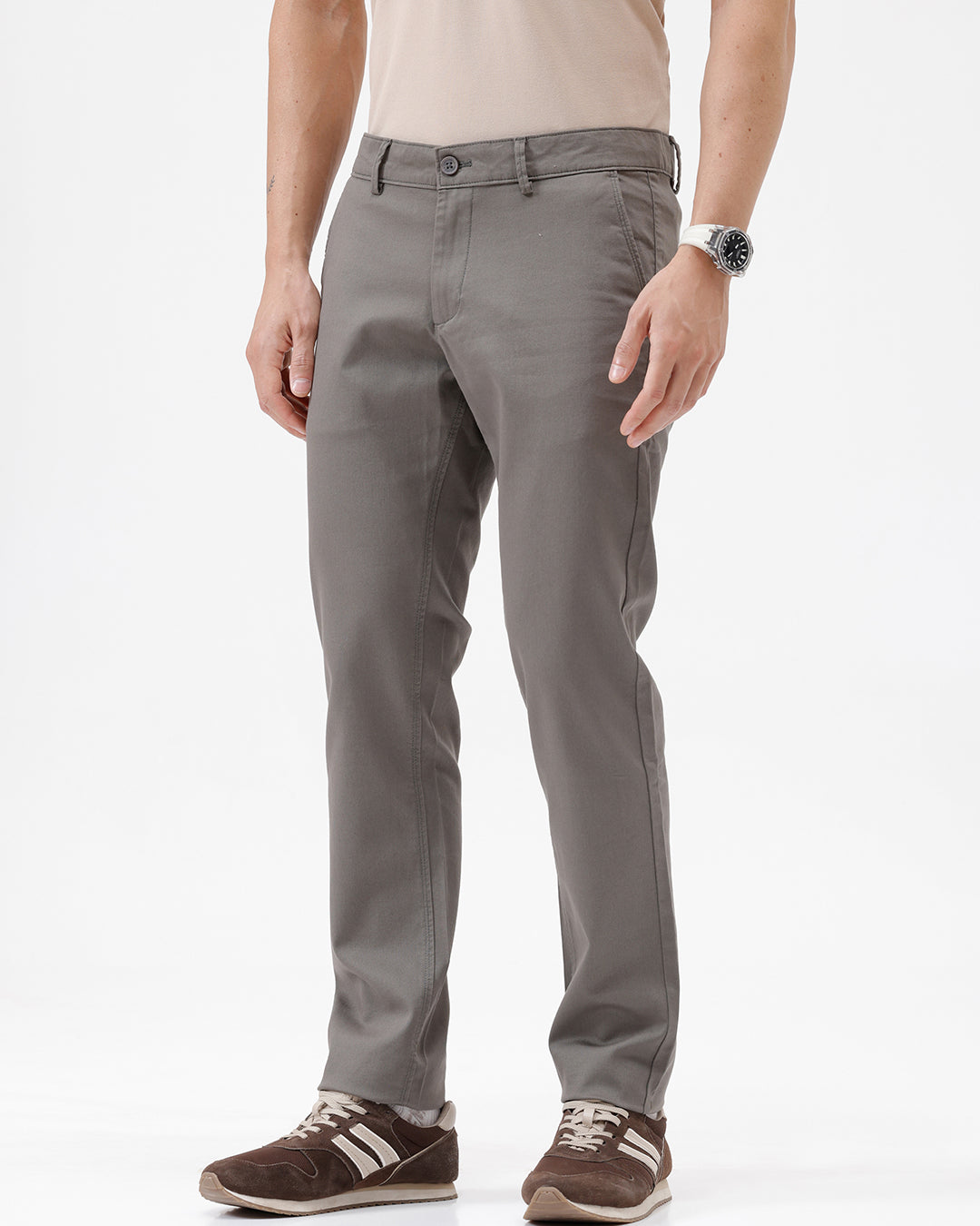 A man wearing slim-fit taupe gray chino pants, paired with a beige t-shirt and brown sneakers, shown from the waist down on a white studio background.