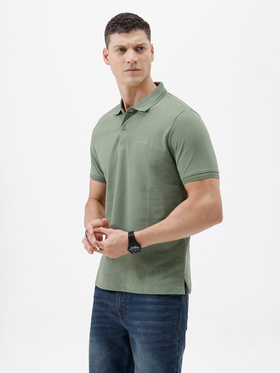 A man wearing a short-sleeve polo shirt in sage green, paired with dark blue jeans, standing against a plain light gray background.