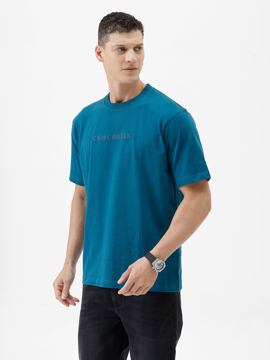 A man wearing a teal blue short-sleeve t-shirt with a subtle tonal 'GREATNESS' text graphic across the chest, paired with black pants, photographed against a clean white studio background.