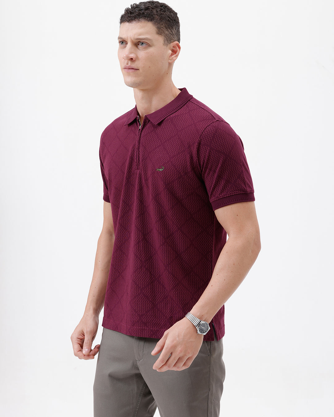 A man wearing a short-sleeve polo shirt in deep burgundy with a subtle textured geometric pattern, styled with gray trousers, sunglasses, and a watch against a plain white background.
