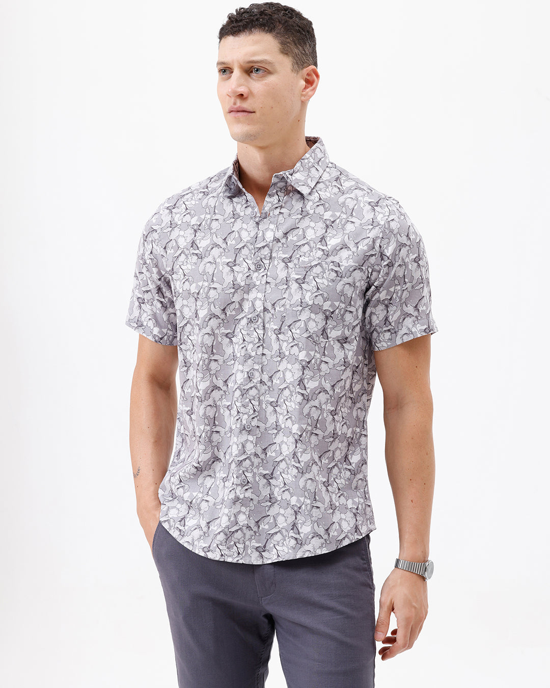 A man wearing a short-sleeve button-up shirt with a gray and white abstract pattern, paired with dark gray trousers, standing against a plain white studio background.