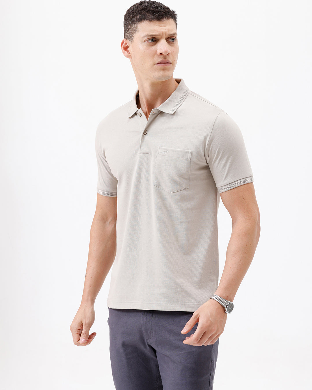 A man wearing a light beige short-sleeve polo shirt with a chest pocket, paired with dark gray trousers, standing against a plain white studio background.