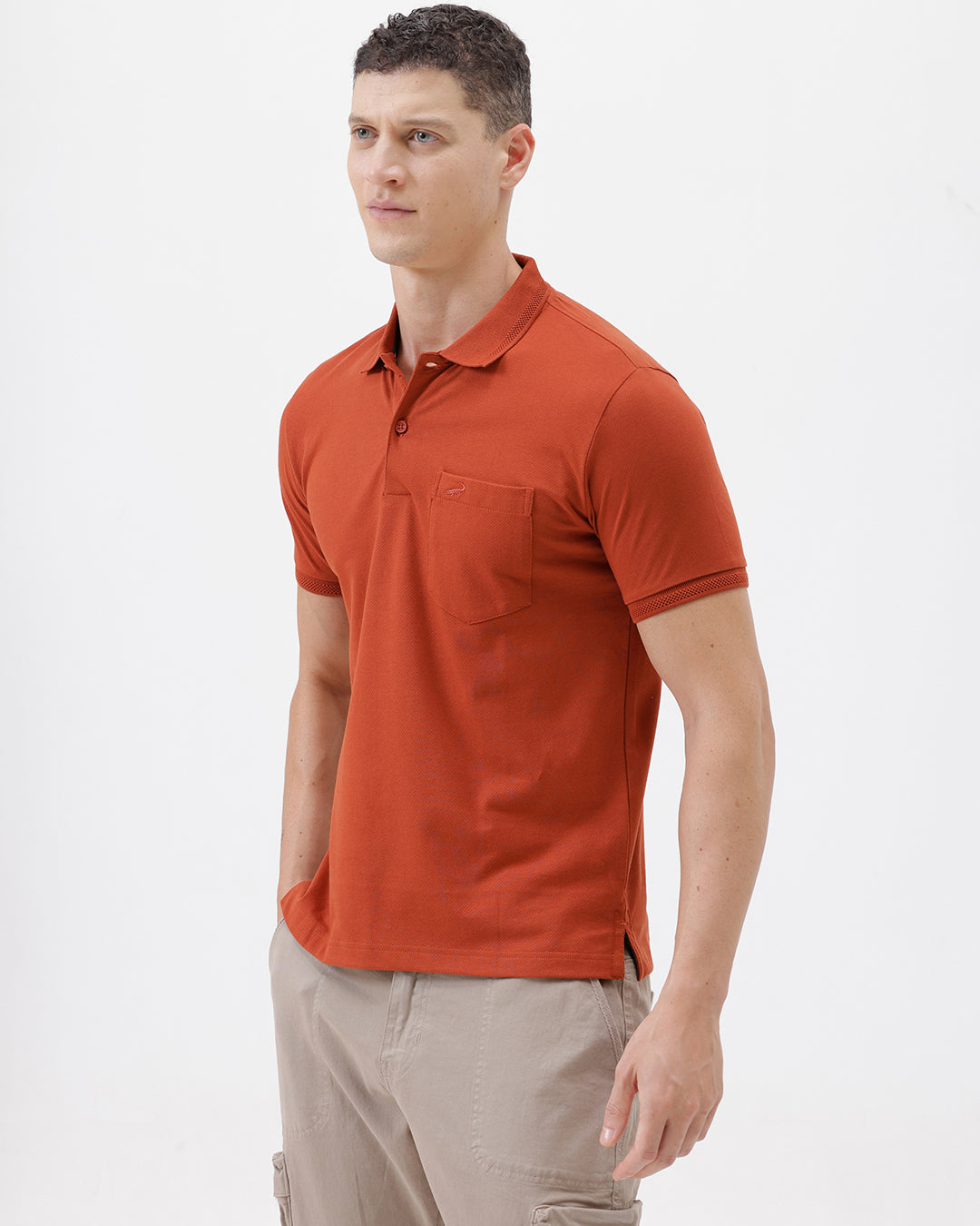 A man wearing a rust-orange short-sleeve polo shirt with a chest pocket, paired with beige cargo pants, standing against a plain light gray studio background.