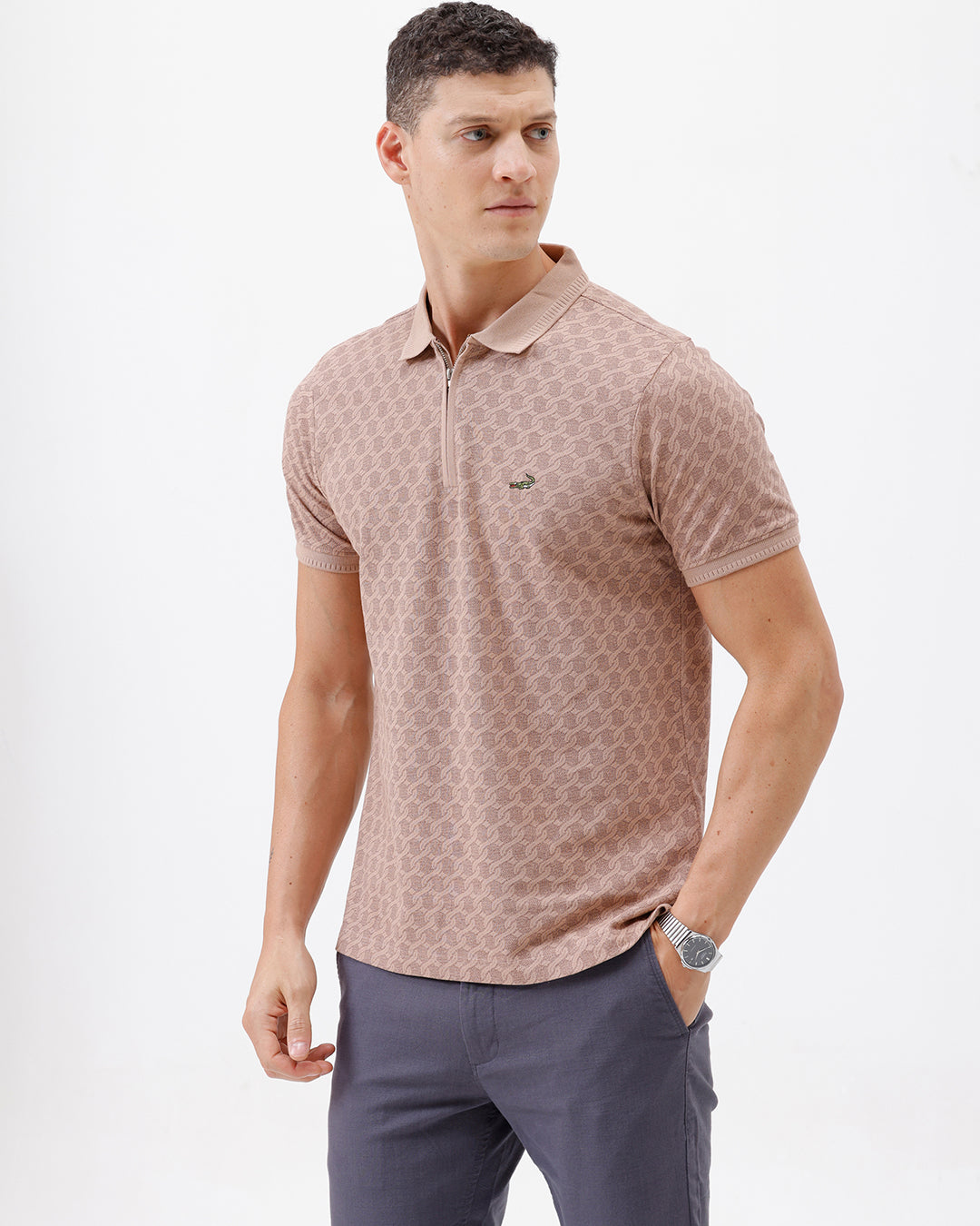 A man wearing a short-sleeve polo shirt with a muted brown and beige geometric pattern, paired with dark gray trousers, standing against a plain white studio background.