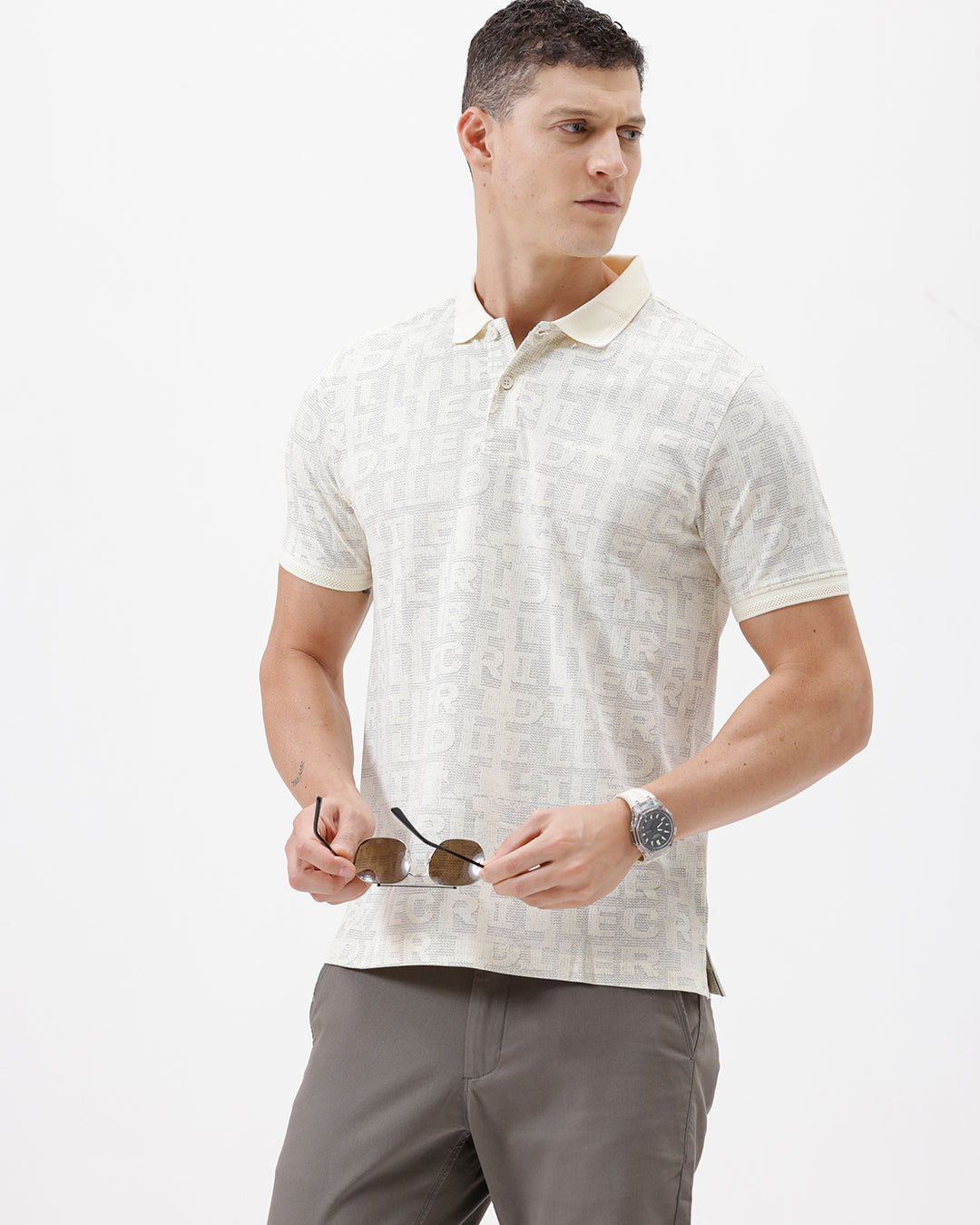 A man wearing a short-sleeve polo shirt with a subtle repeating monogram pattern in cream and light gray, paired with gray trousers, photographed against a plain white studio background.