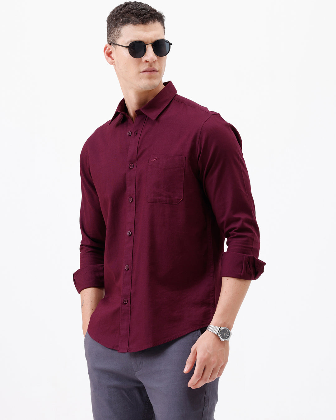 A man wearing a long-sleeve button-down shirt in deep burgundy with rolled-up sleeves and gray trousers, standing against a plain white studio background.
