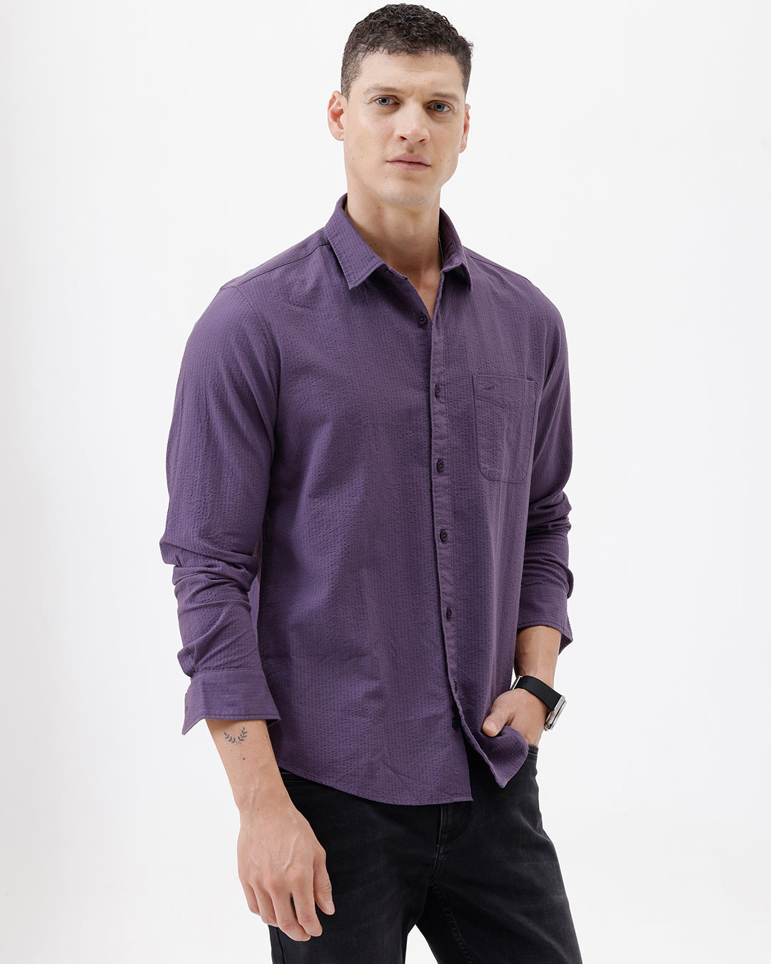 A man wearing a long-sleeve button-down shirt in a muted purple color, paired with black pants, standing against a plain white studio background.