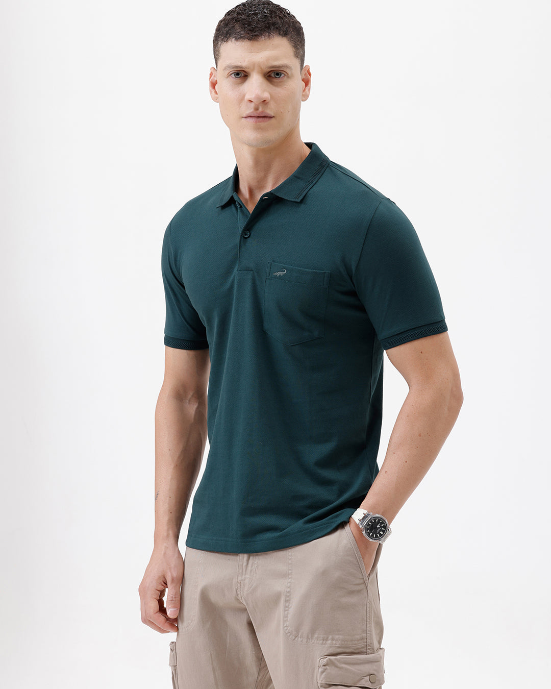 A man wearing a dark teal short-sleeve polo shirt with a chest pocket, paired with beige cargo pants, standing against a plain white studio background.