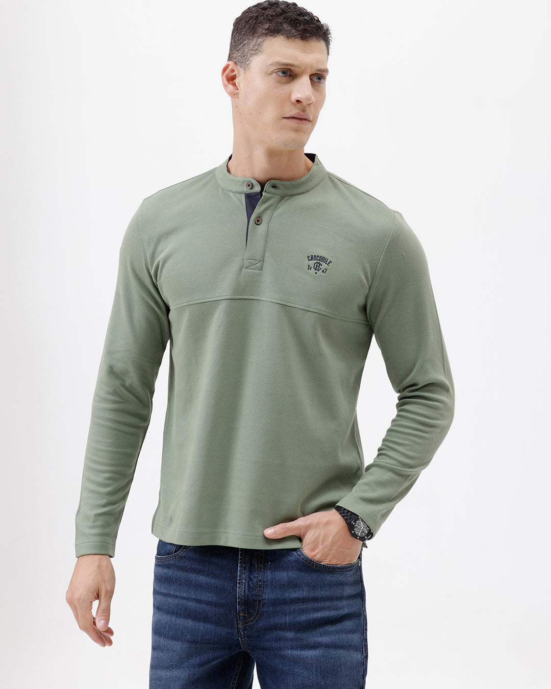 A man wearing a sage green long-sleeve henley shirt with a small embroidered logo on the left chest, paired with blue jeans, against a plain white studio background.