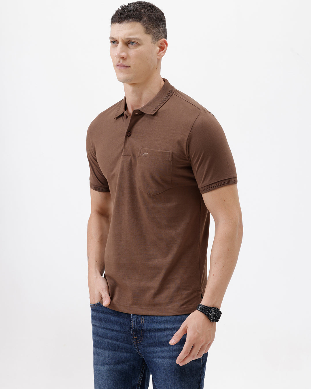 A man wearing a brown short-sleeve polo shirt with a chest pocket, paired with blue jeans, standing against a plain light gray background.