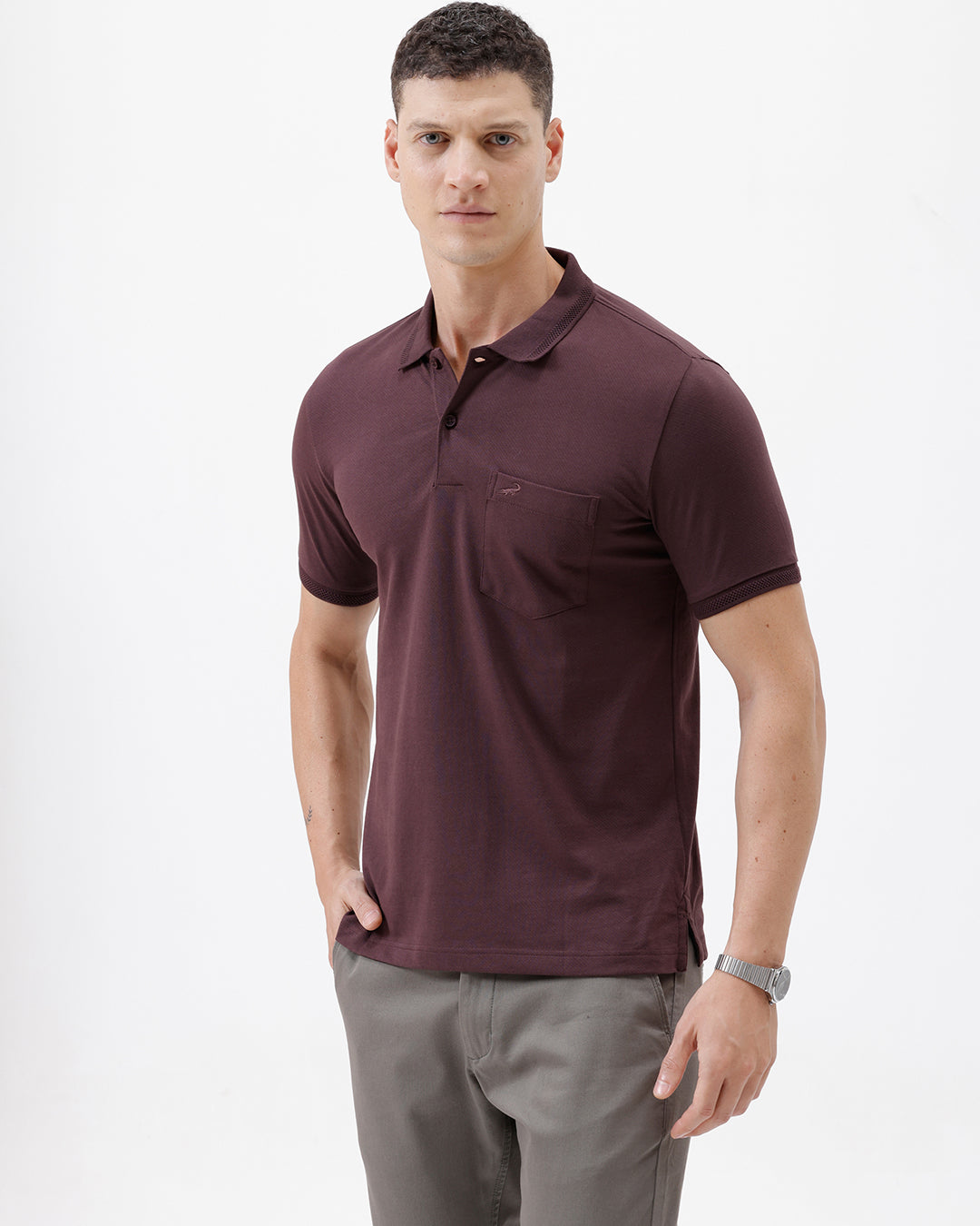 A man wearing a short-sleeve polo shirt in dark burgundy with a chest pocket, paired with gray trousers, standing against a plain light gray background.