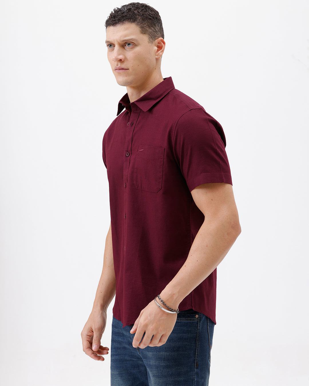 A man wearing a short-sleeve button-up shirt in maroon with a chest pocket, paired with blue jeans, standing against a plain white studio background.