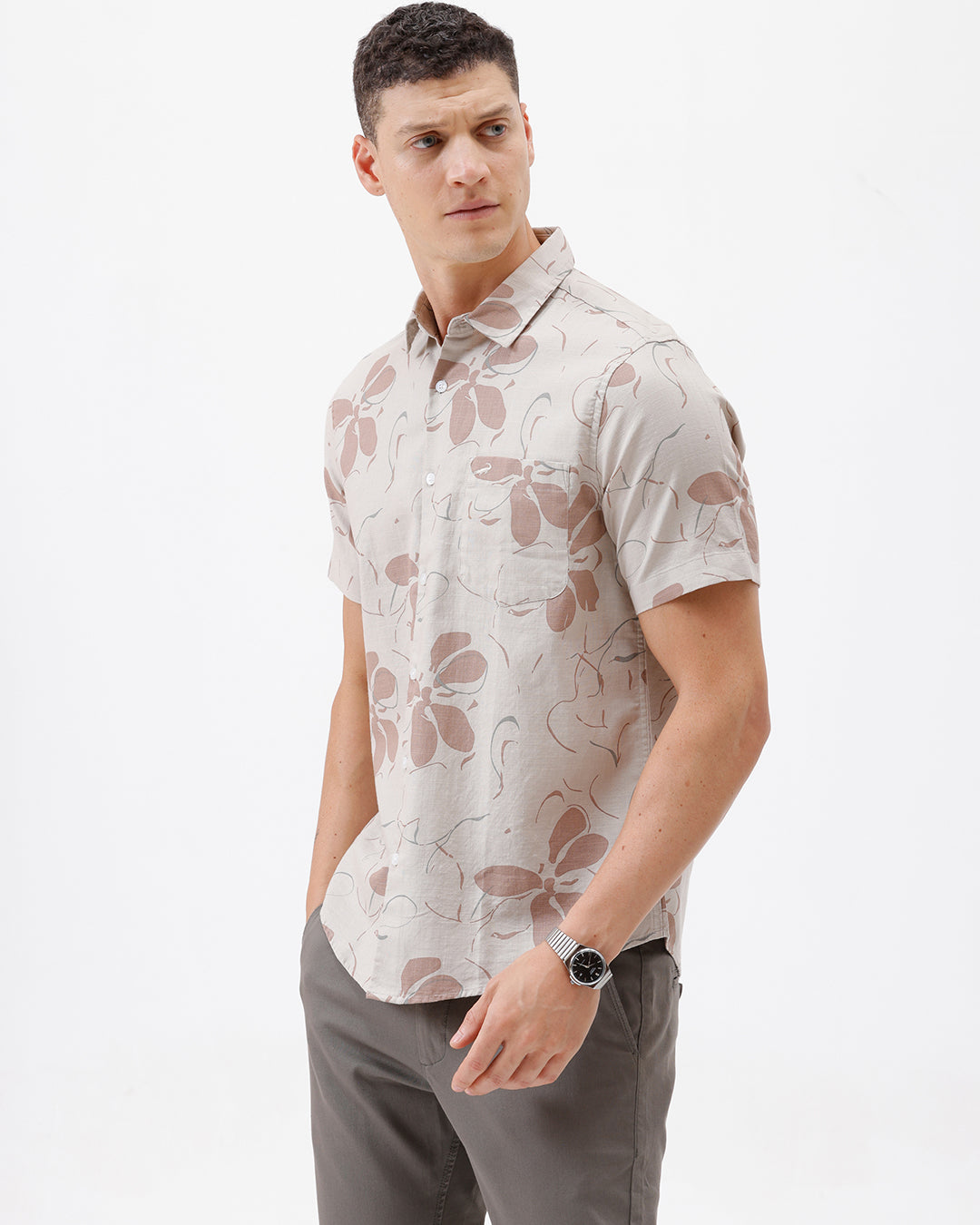 A man wearing a short-sleeve button-up shirt with a beige and brown floral pattern, paired with gray trousers, standing against a plain white background.