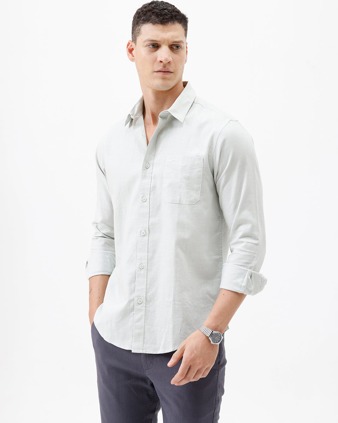 A man wearing a light sage green long-sleeve button-down shirt with rolled-up sleeves and dark gray trousers, standing against a plain white studio background.