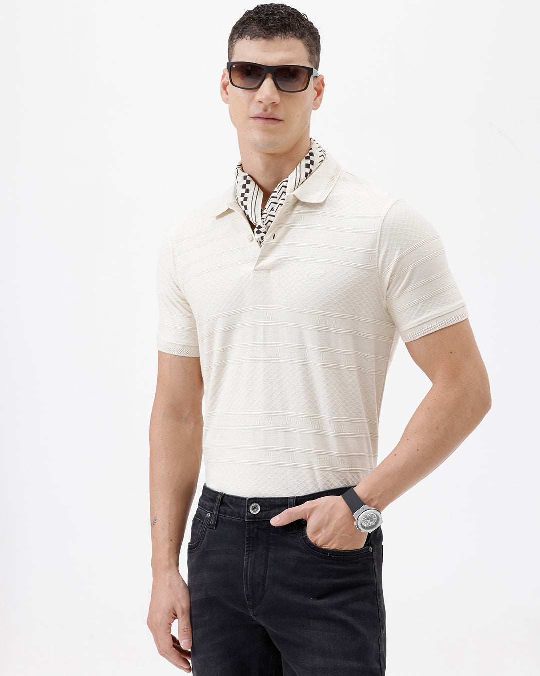 A man wearing a short-sleeve polo shirt in cream with subtle horizontal textured stripes, paired with dark jeans, standing against a plain white studio background.