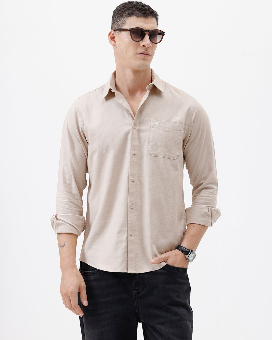 A man wearing a light beige long-sleeve button-up shirt with a chest pocket, paired with dark jeans, standing against a plain white studio background.