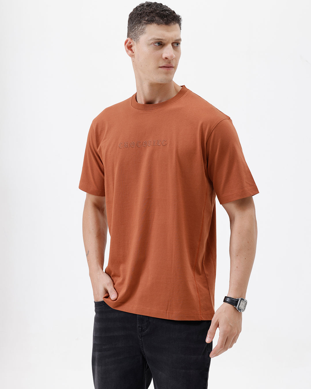 A man wearing a rust-orange crewneck t-shirt with a subtle tonal 'ELECTRIC' logo across the chest, paired with black pants, photographed against a clean white studio background.