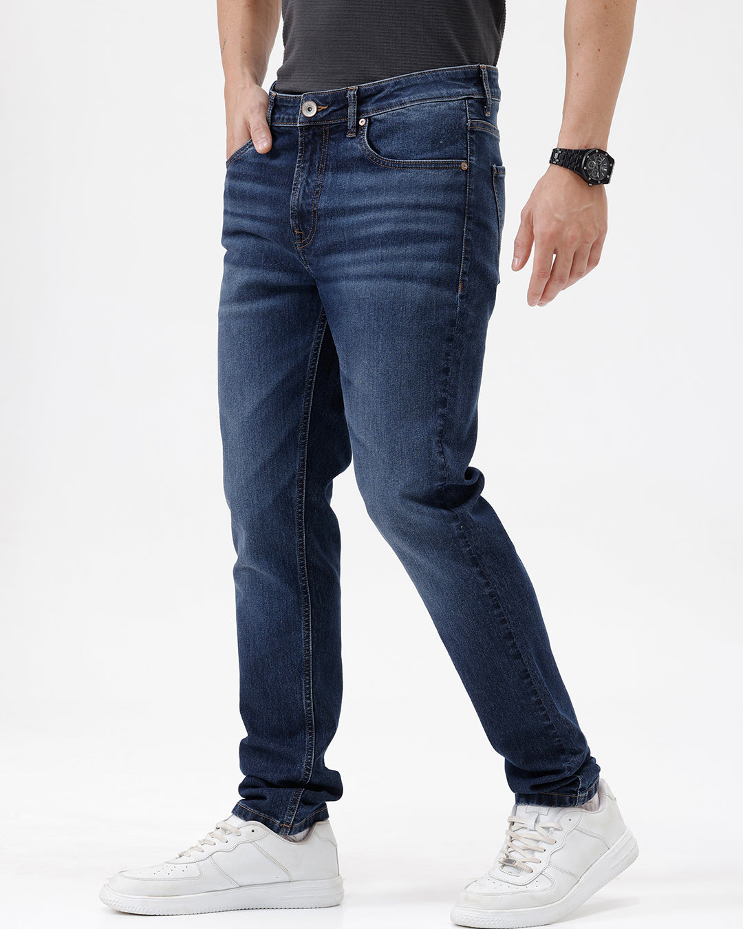 A person wearing dark blue slim-fit jeans with a faded wash, paired with a black t-shirt and white sneakers, shown from the waist down against a plain white studio background.