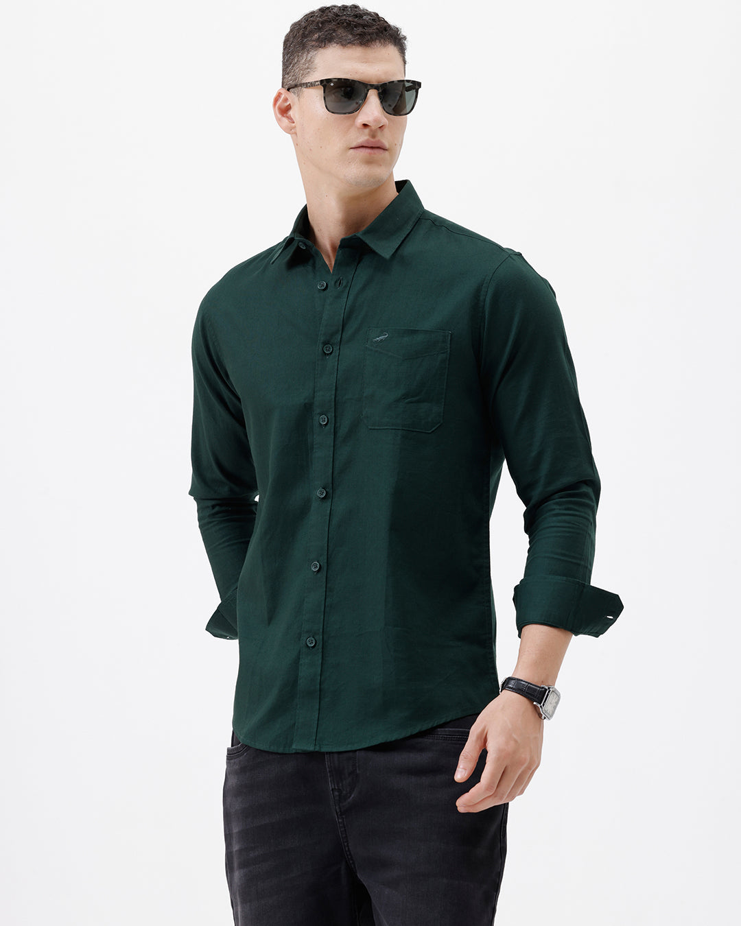 A man wearing a dark forest green long-sleeve button-down shirt with a chest pocket, paired with dark jeans, standing against a plain white studio background.