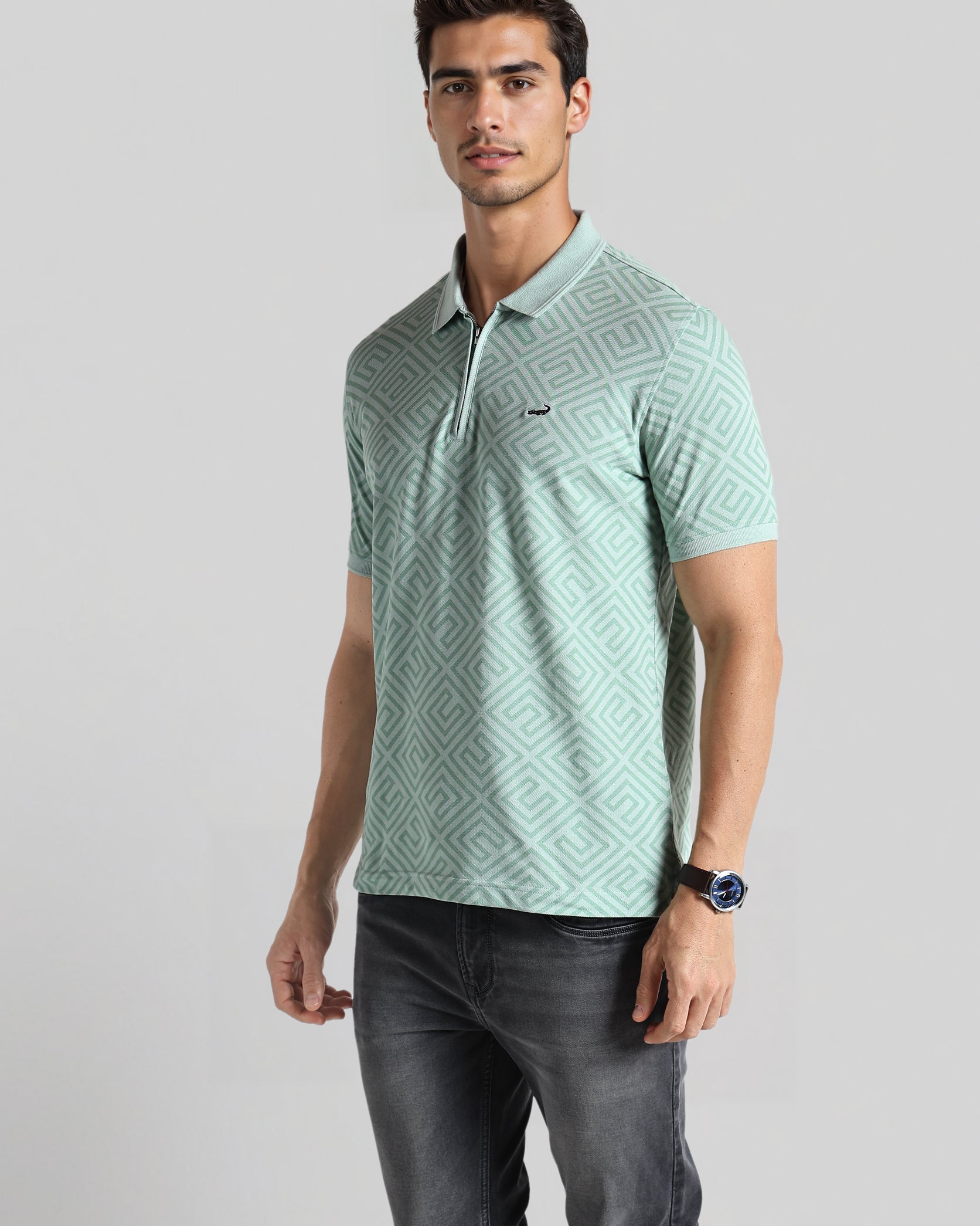 Man wearing a light green patterned polo shirt and dark jeans on a gray background