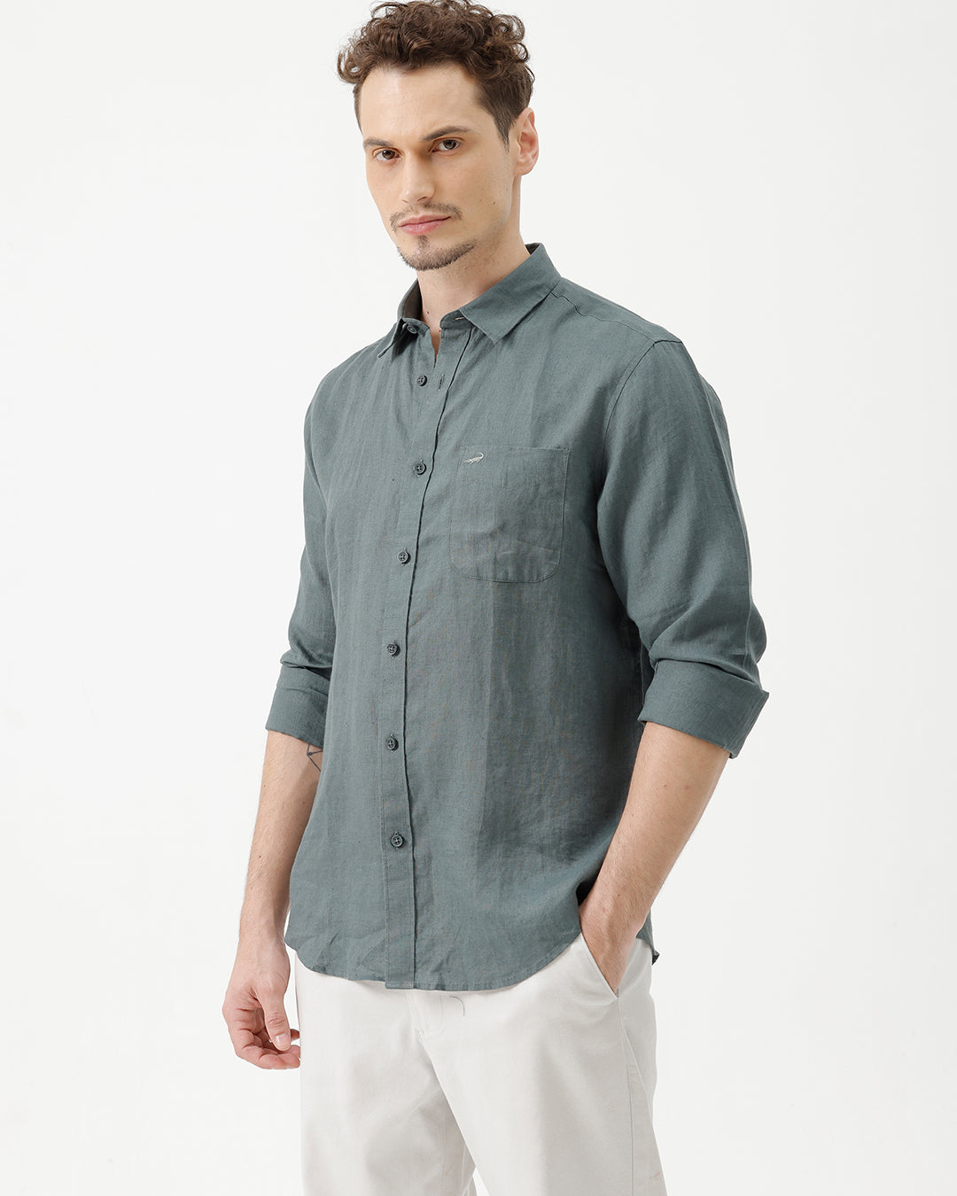 A man wearing a slate blue-green long-sleeve linen button-up shirt with white trousers, standing against a plain white studio background.