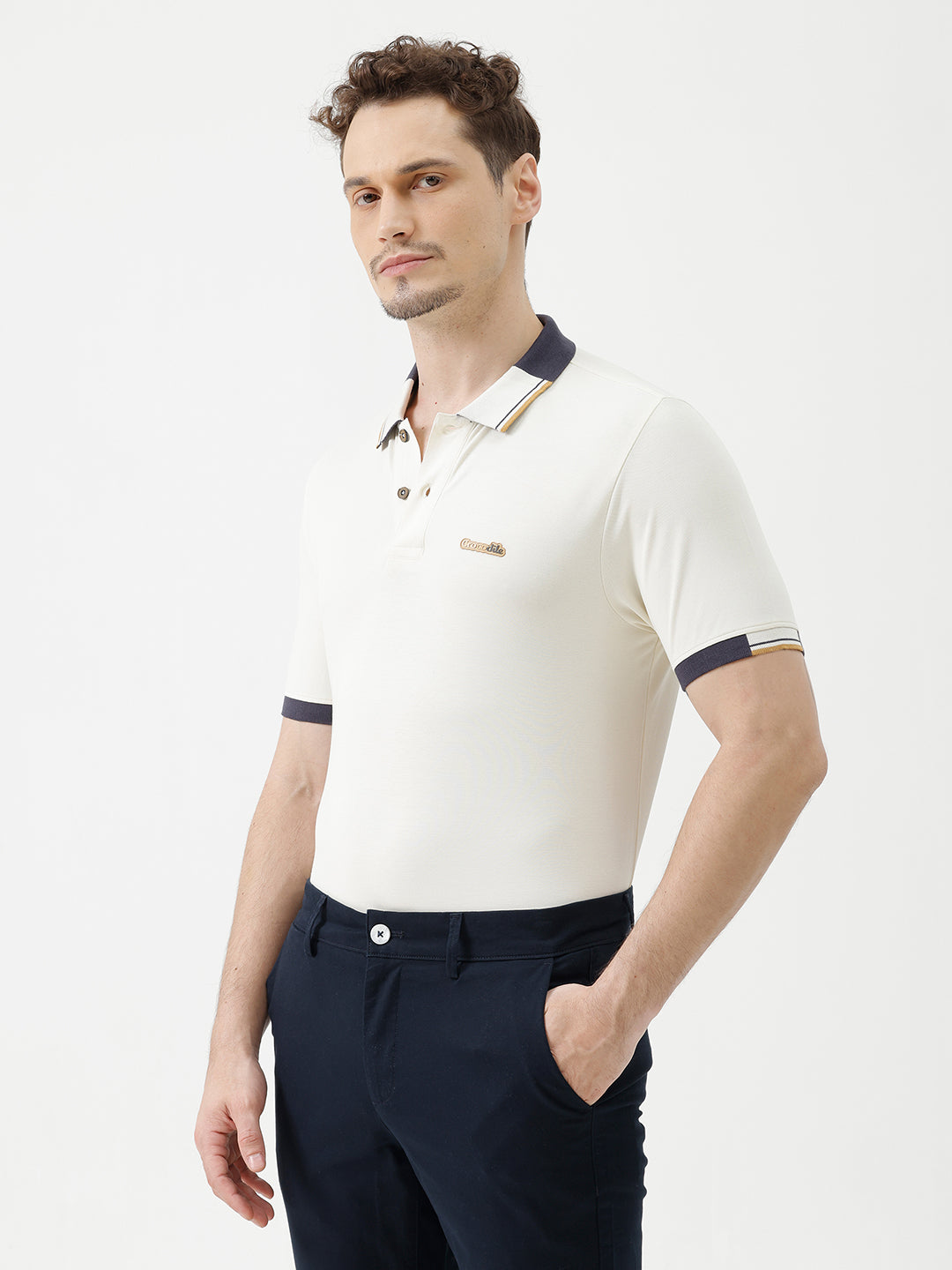 A man wearing a short-sleeve polo shirt in off-white with navy blue and gold trim on the collar and sleeve cuffs, paired with dark navy trousers, against a plain light gray background.