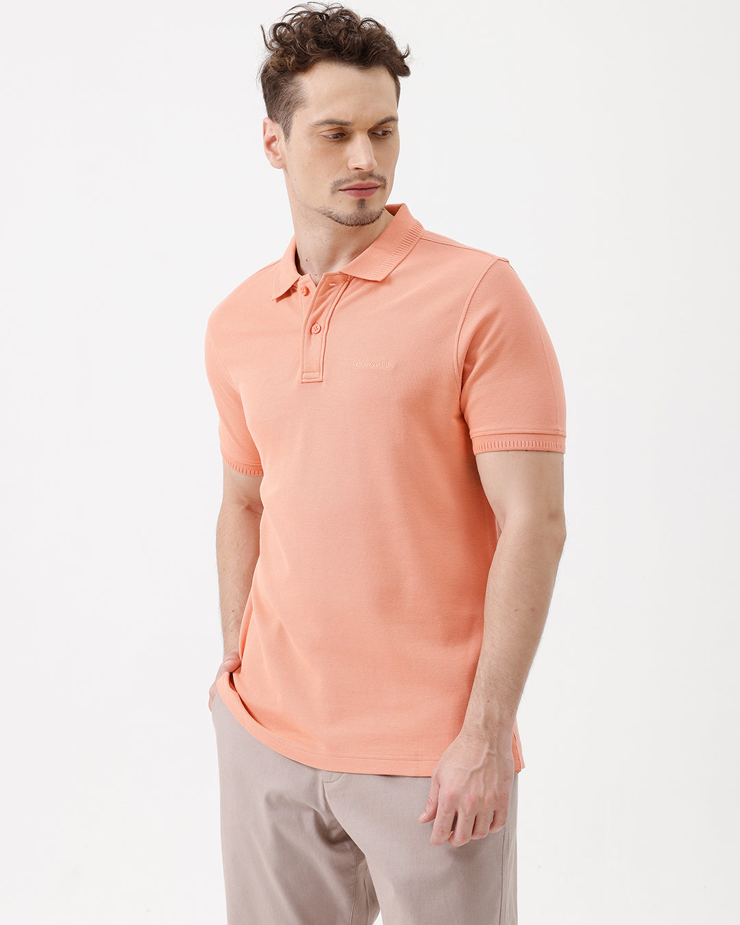 A man wearing a peach-colored short-sleeve polo shirt and beige trousers, standing against a plain white studio background.