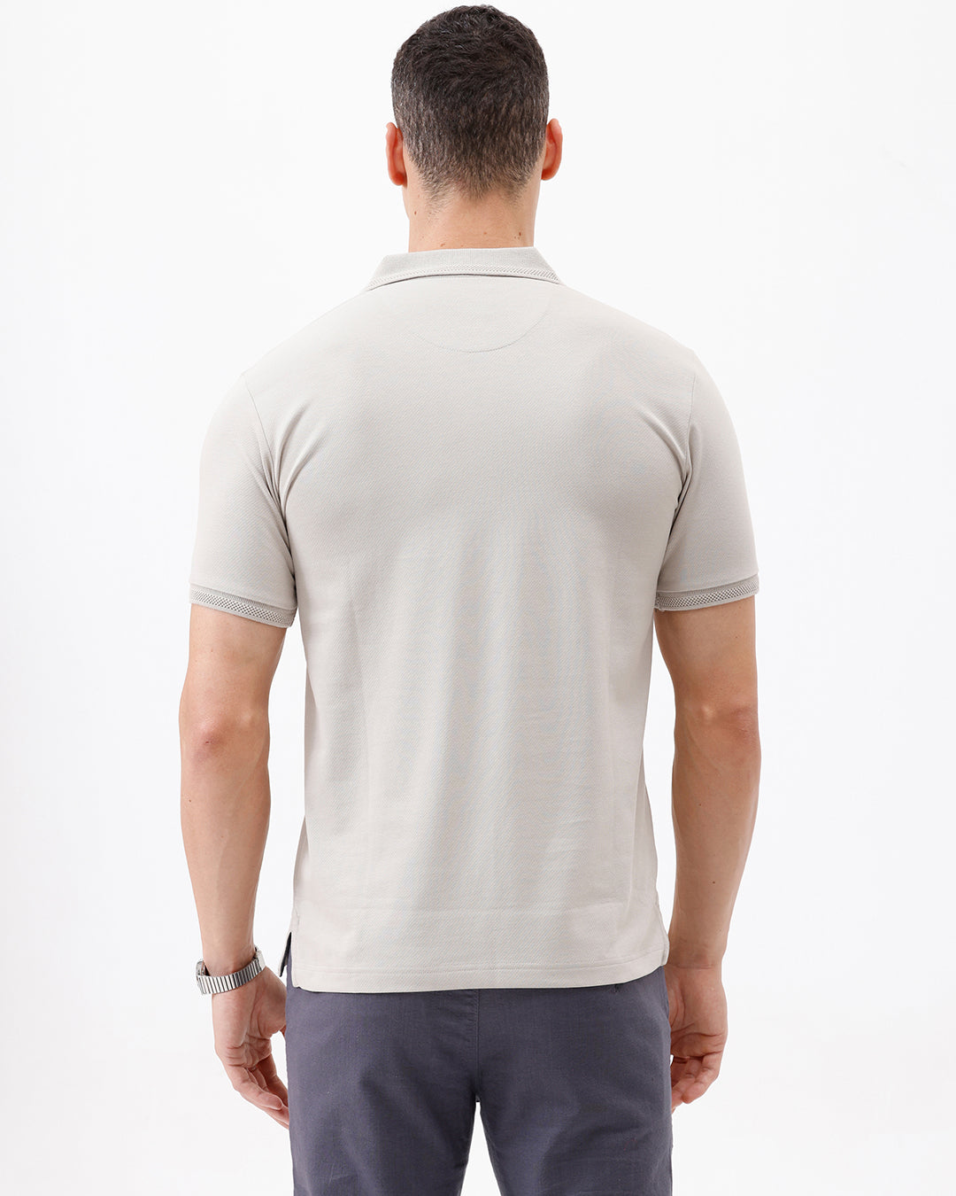 A man wearing a light beige short-sleeve polo shirt with a chest pocket, paired with dark gray trousers, standing against a plain white studio background.
