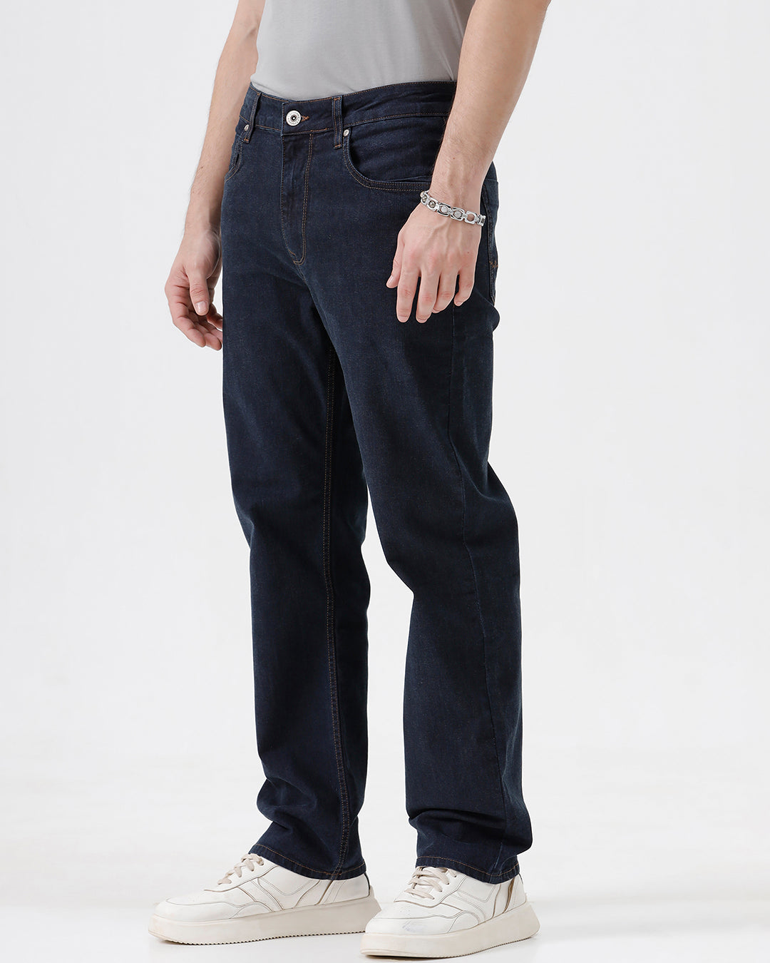 A person wearing dark navy blue straight-leg jeans, a light gray t-shirt, and white sneakers, standing against a plain white studio background.
