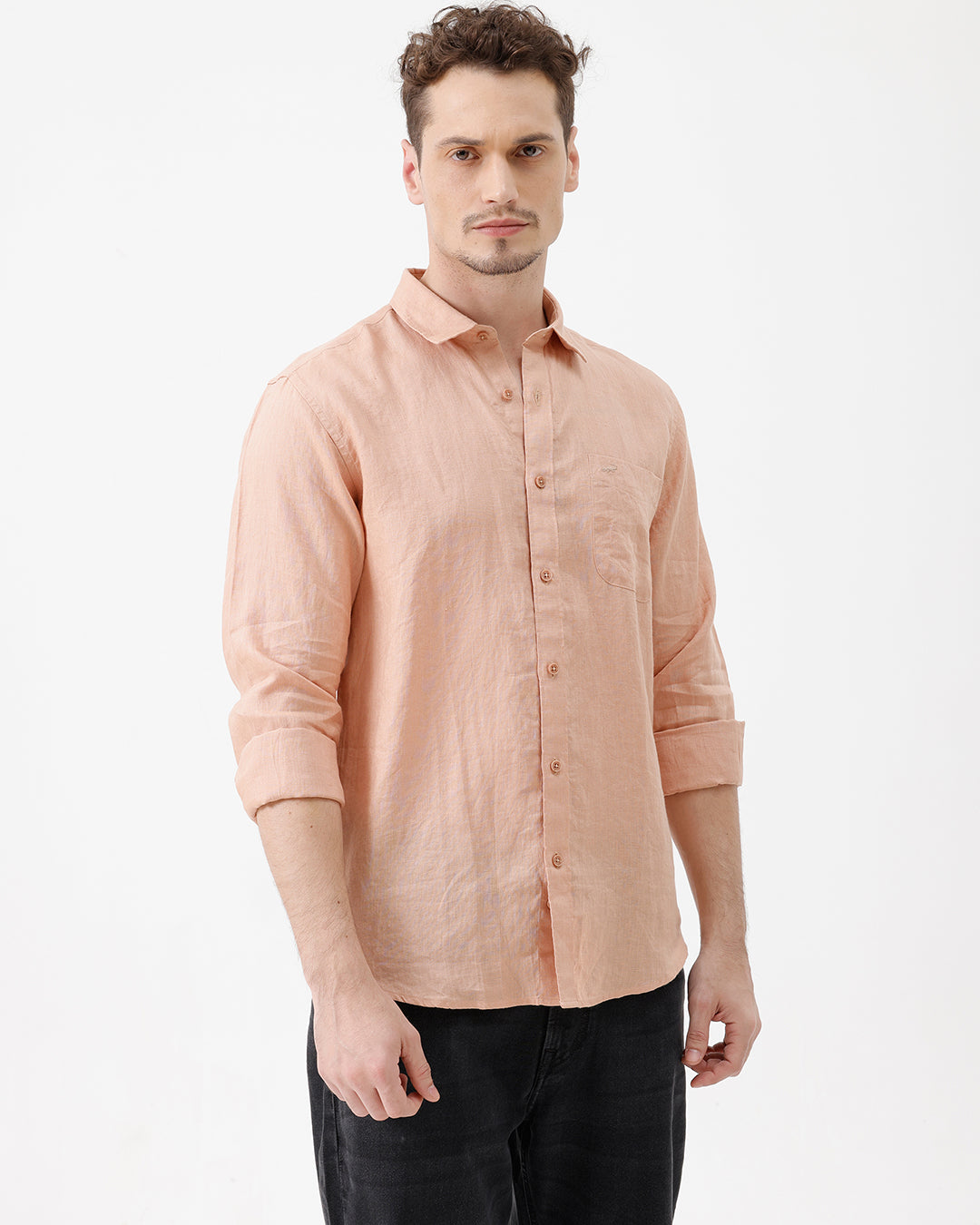 A man wearing a light peach-colored linen button-down shirt with dark jeans, standing against a plain white studio background.