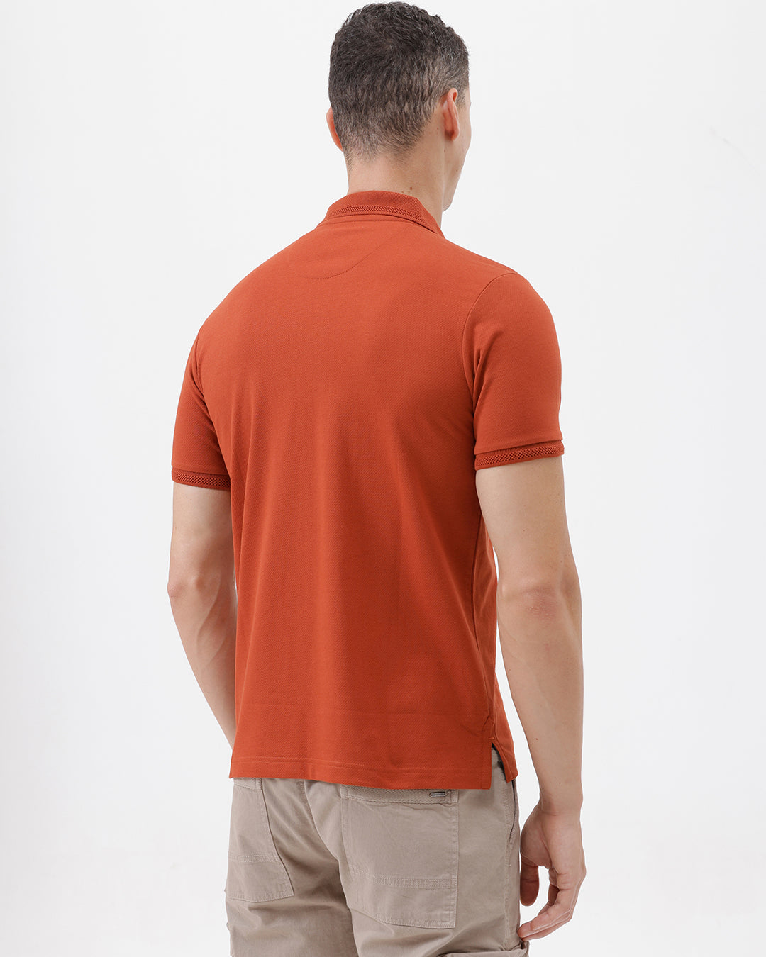 A man wearing a rust-orange short-sleeve polo shirt with a chest pocket, paired with beige cargo pants, standing against a plain light gray studio background.